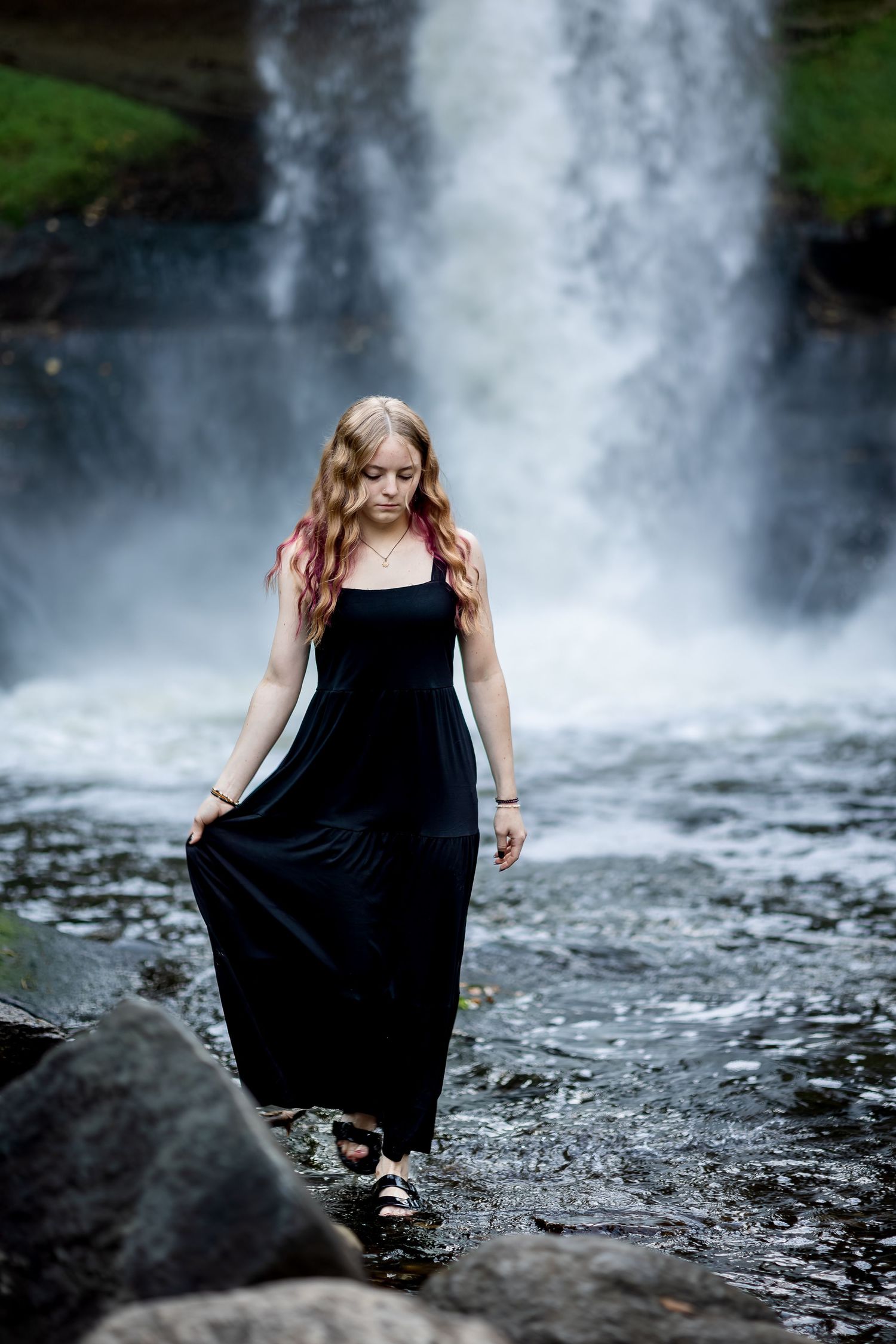 A person in a flowing black dress poses gracefully near a misty waterfall on rocky terrain.