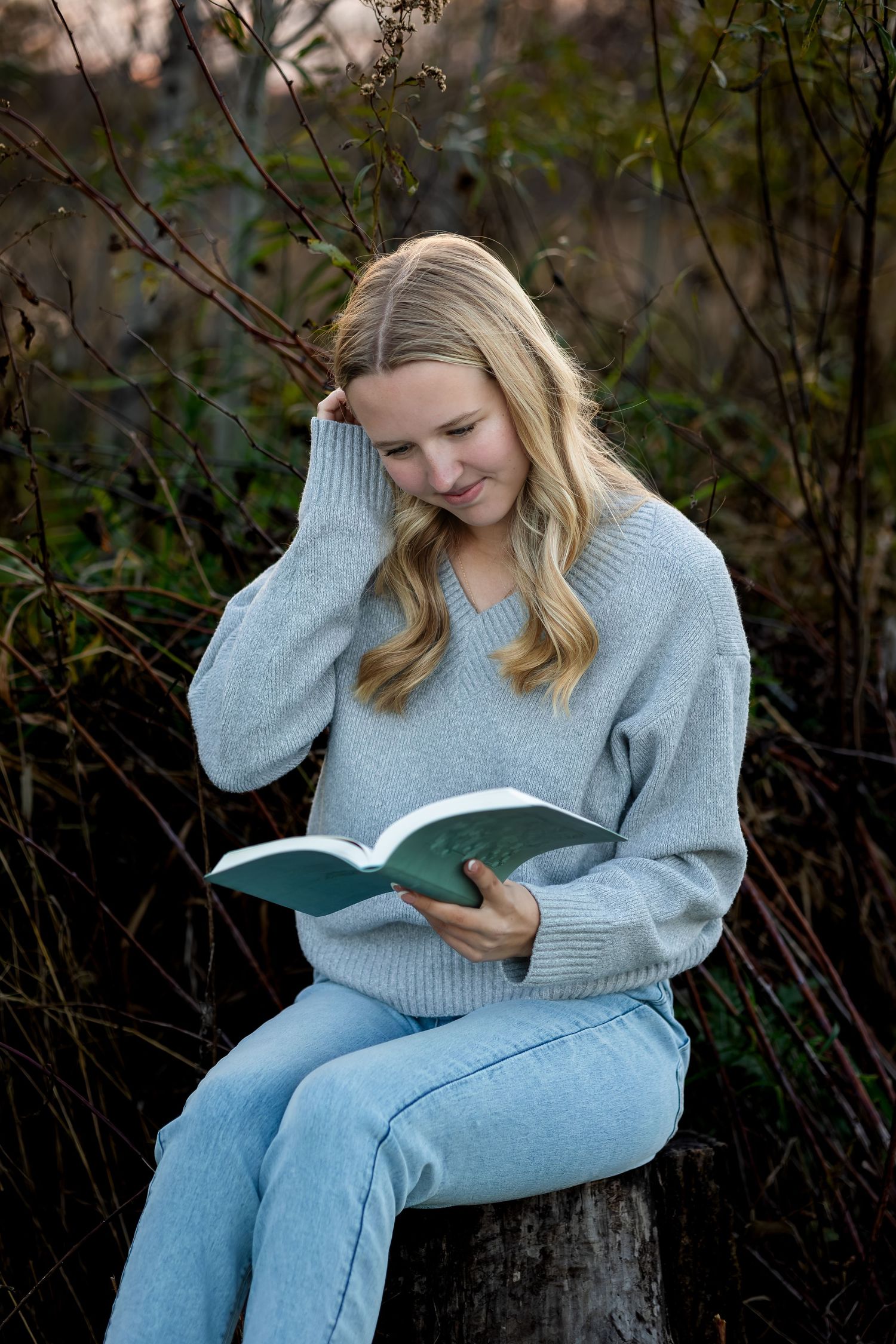 A person in a gray sweater and jeans sits on a log while reading a book outdoors.