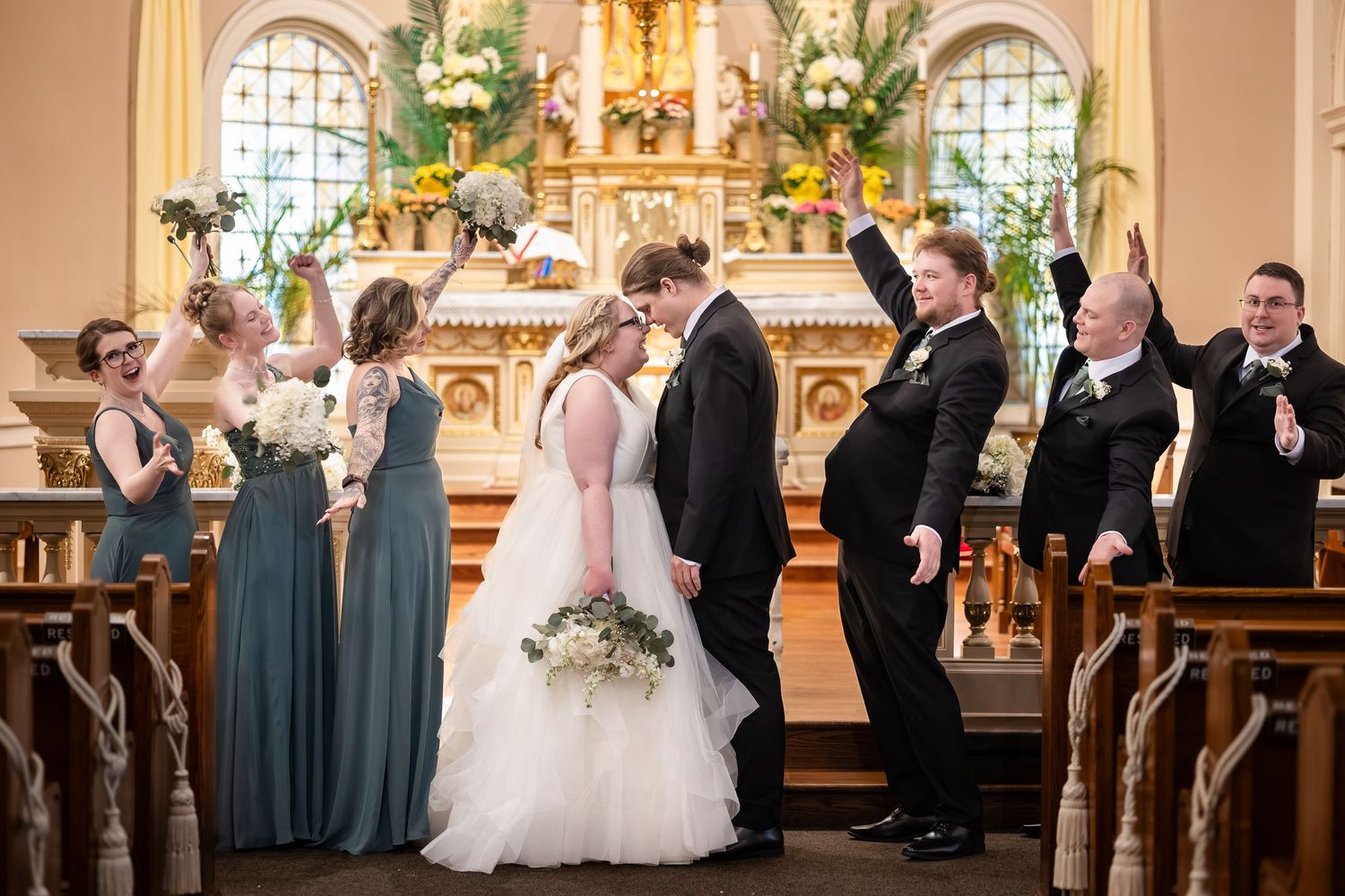 Bride and groom share first kiss at church altar surrounded by bridesmaids in dusty blue gowns and cheering groomsmen.