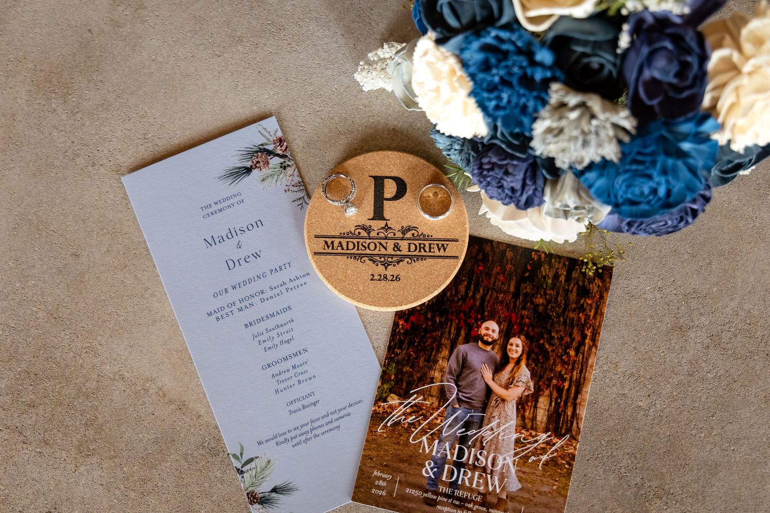 Wedding stationery, program, personalized wooden coaster, and blue floral bouquet arranged on a surface.