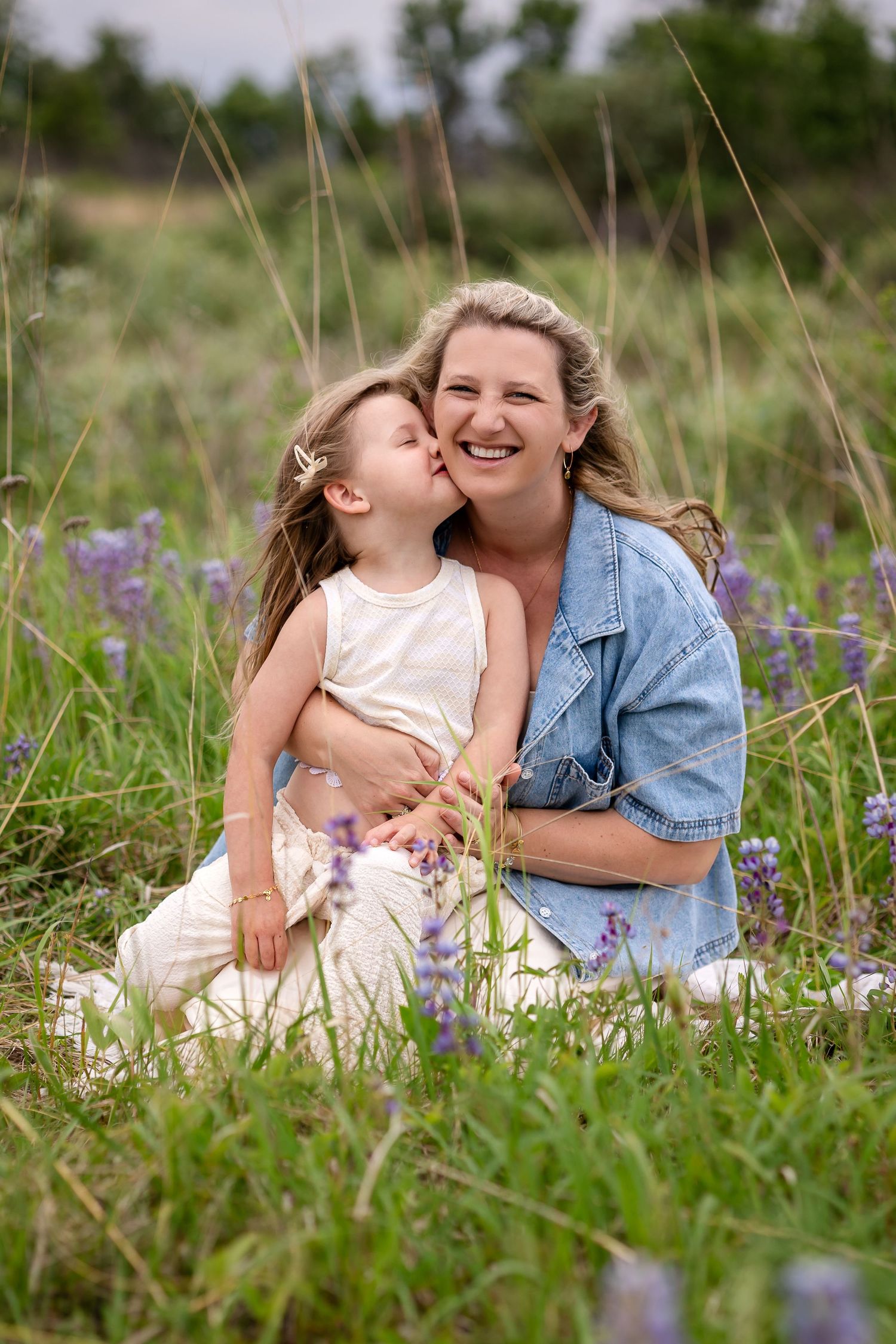 A young girl kisses her smiling mother while sitting together in a field of purple wildflowers.