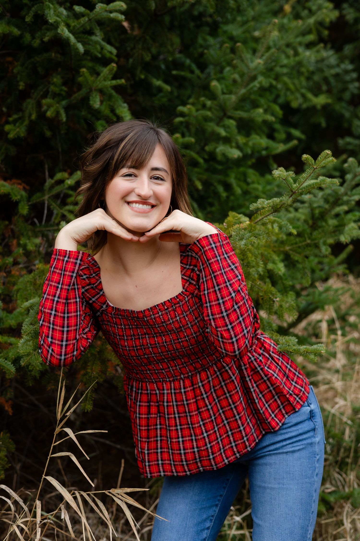Person in red plaid top and jeans poses playfully outdoors with trees in background.