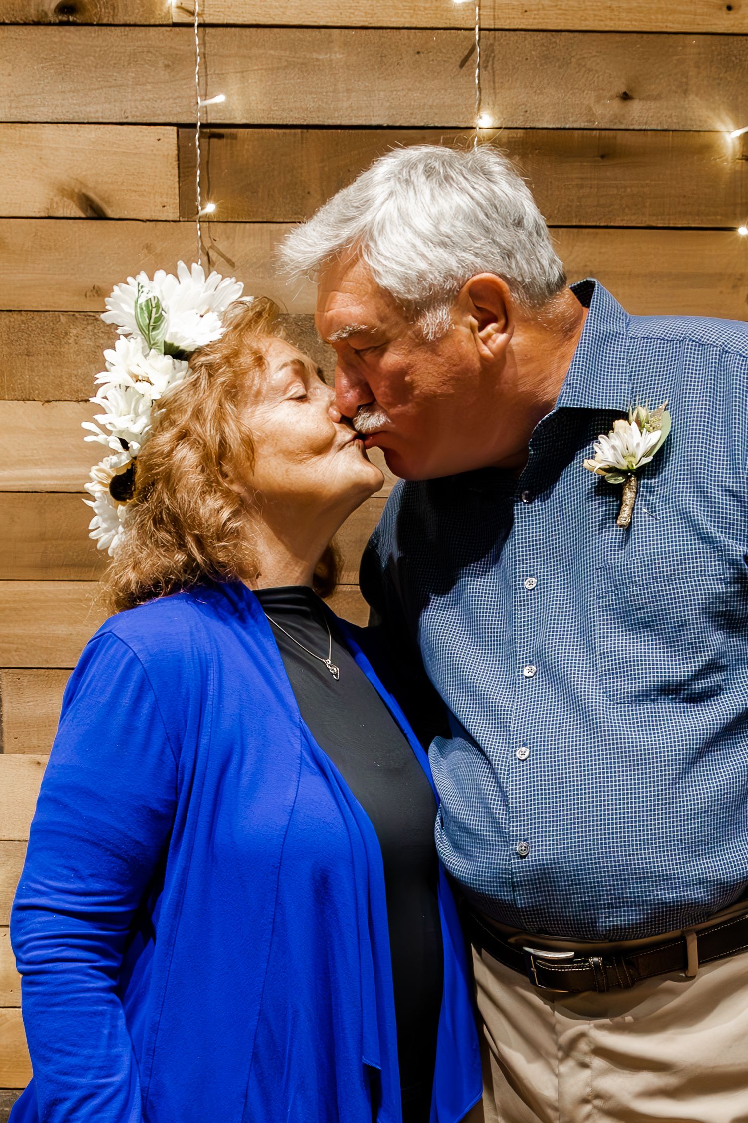 A romantic moment between a couple wearing blue and white floral accessories against a wooden wall with string lights.