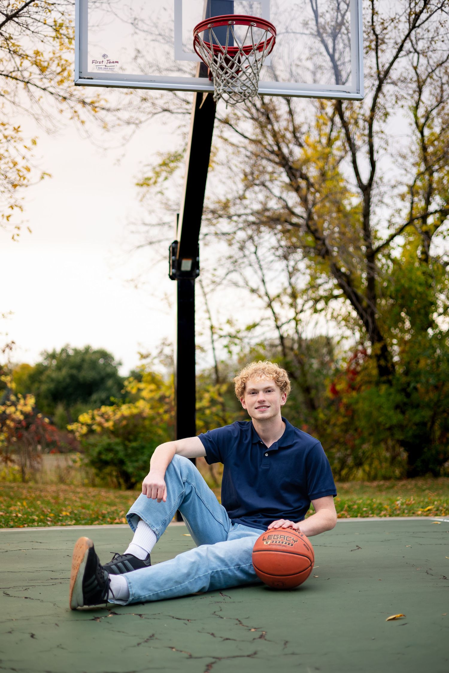 Basketball player relaxes on outdoor court with ball next to hoop during autumn season.