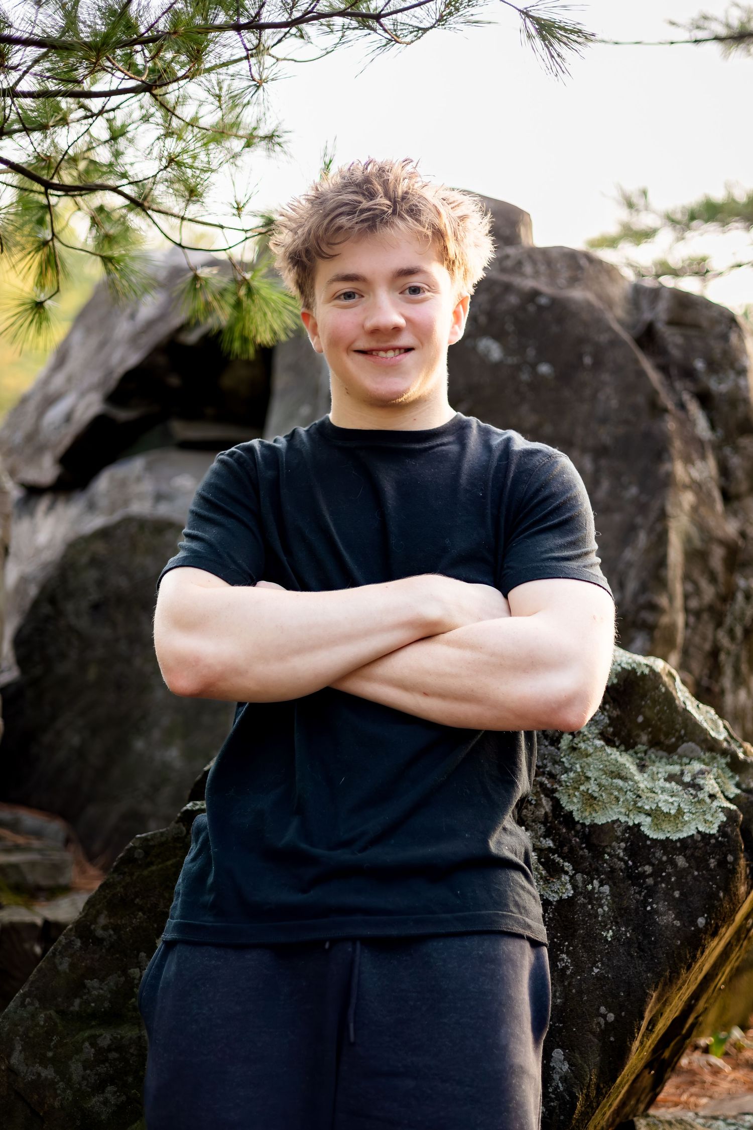 A young person in a navy t-shirt stands with crossed arms in front of large rocks and pine branches outdoors.