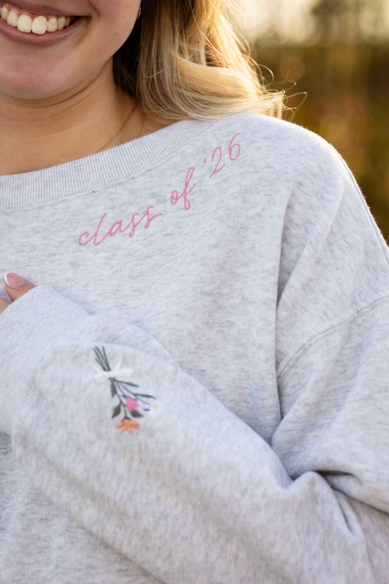 A close-up of a gray sweatshirt with 'class of 26' text and a small floral design embroidered on it.