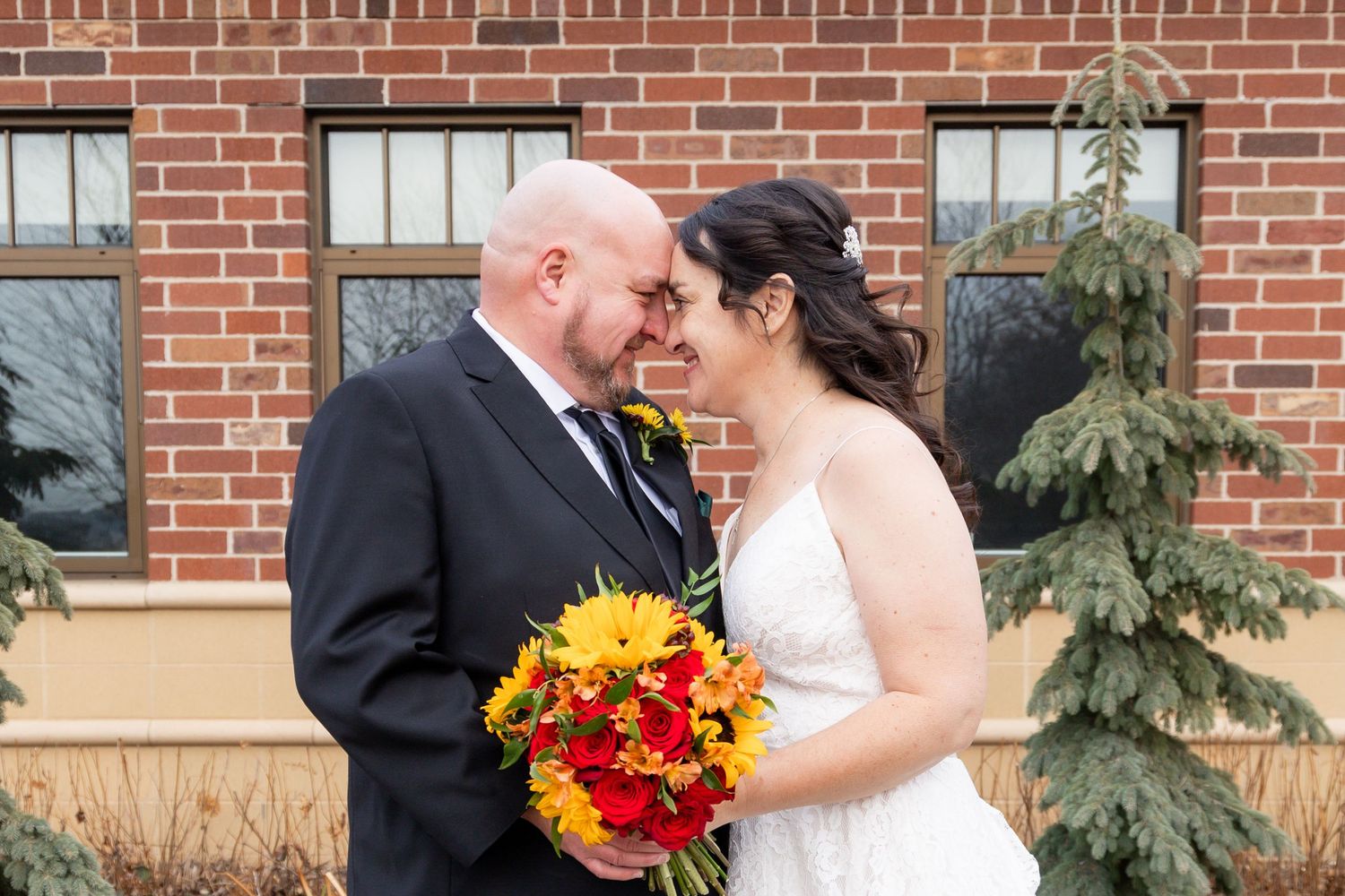A bride and groom share an intimate moment against a brick building with fall-colored flowers.