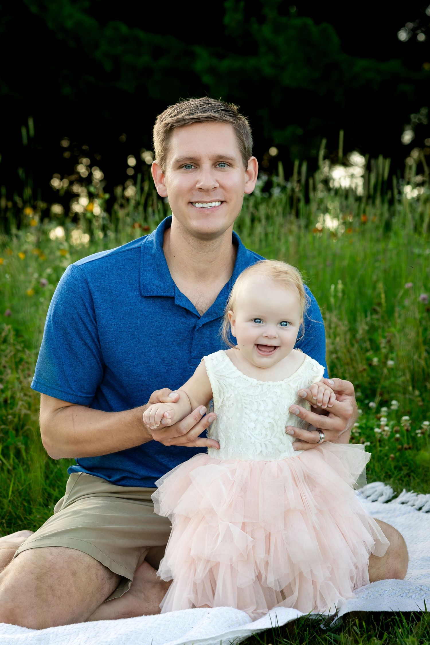 Parent and baby in pink dress share joyful moment on blanket outdoors.