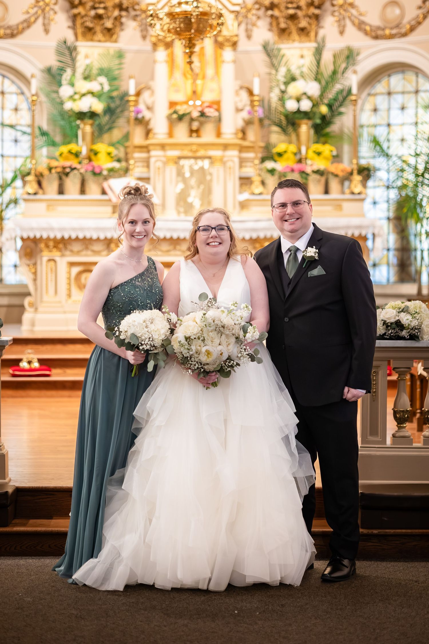 Bride in white ruffled gown poses with bridesmaid and groomsman at church altar.