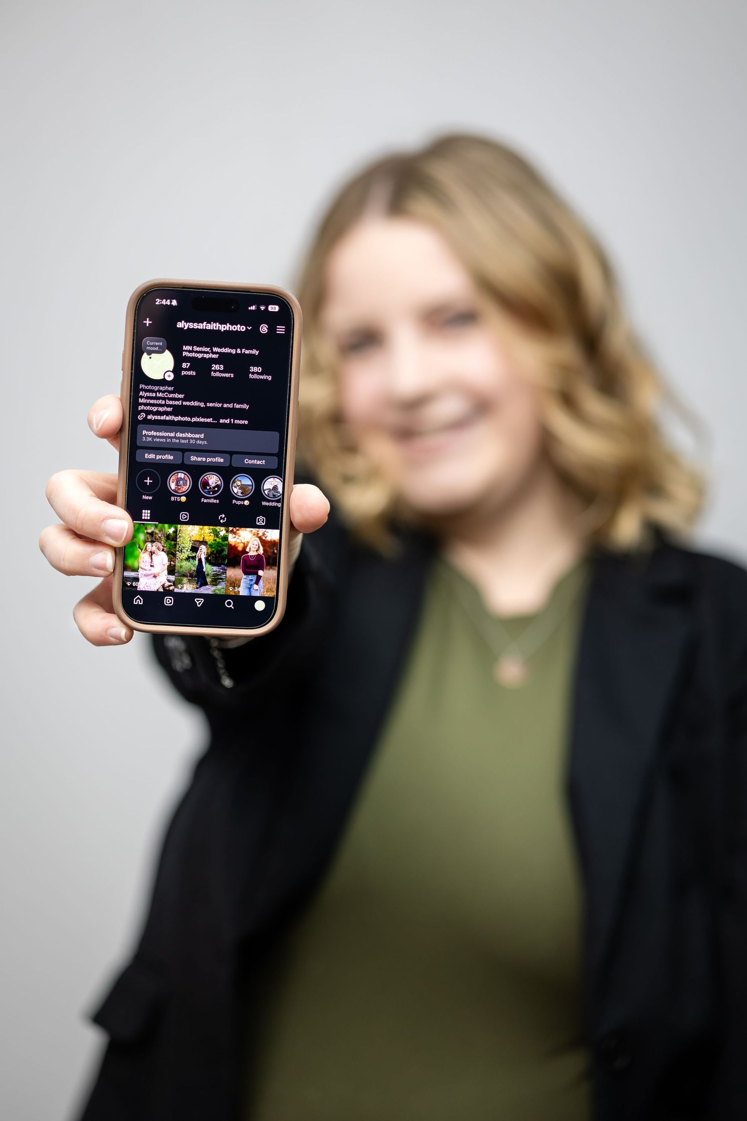 A person wearing a green top and black blazer holds up a smartphone displaying a dark-themed social media app interface.