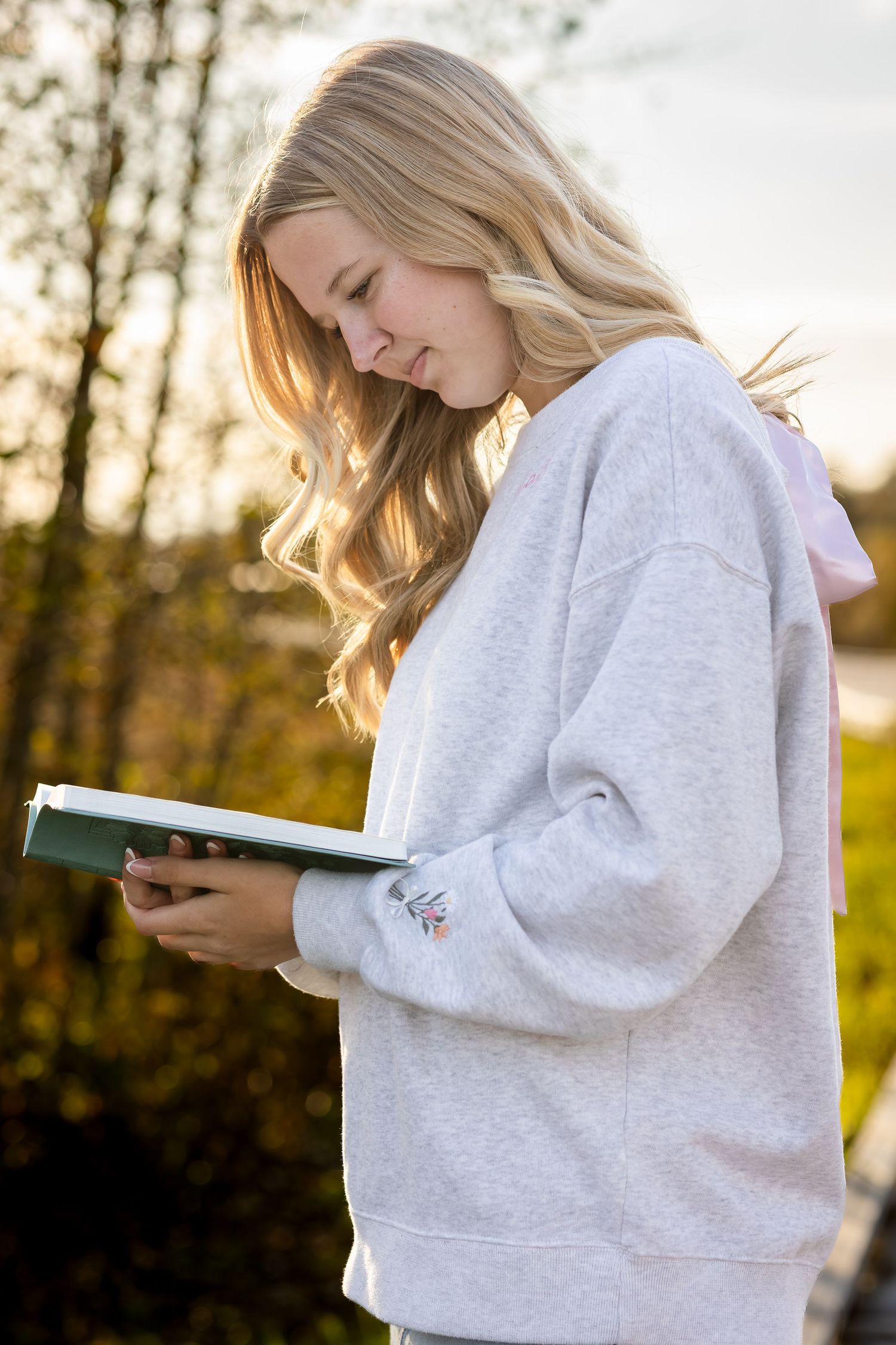 Person in a light gray sweatshirt reading on a mobile device outdoors during golden hour with trees in background.