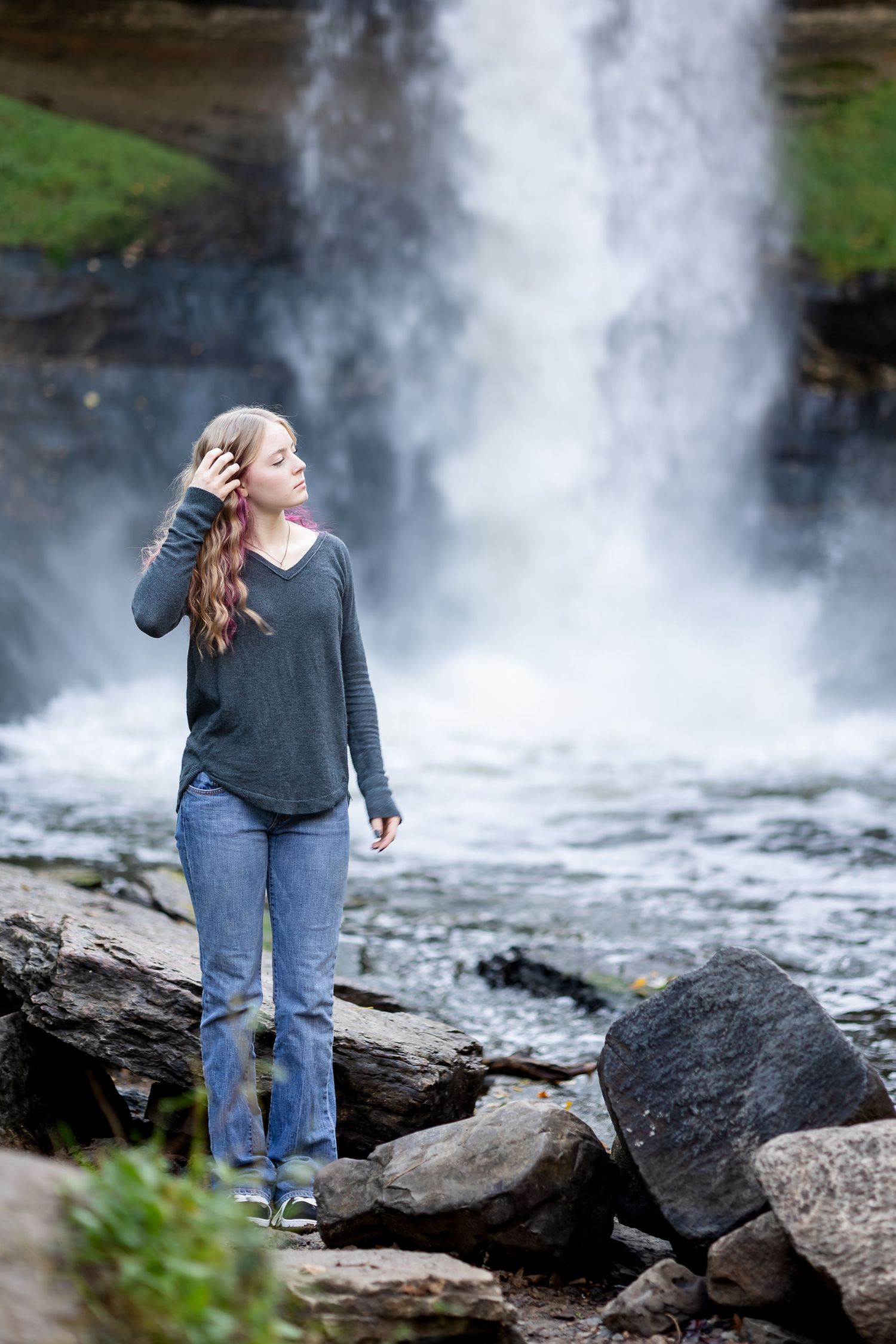 A person stands on dark rocky terrain near a misty waterfall in a scenic natural setting.