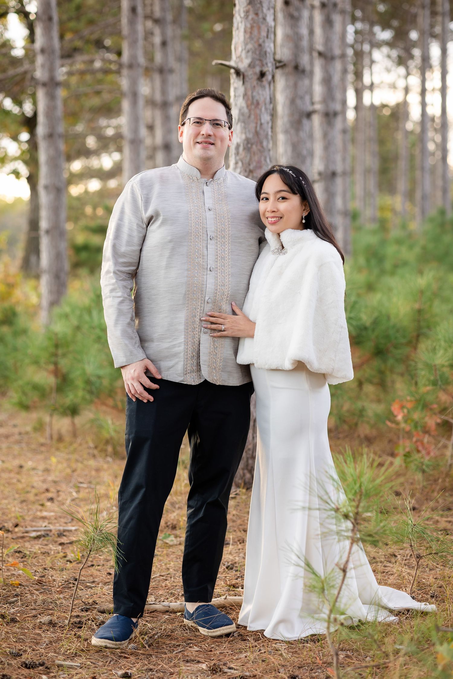 A couple in formal attire poses among pine trees, with bride wearing elegant white dress.