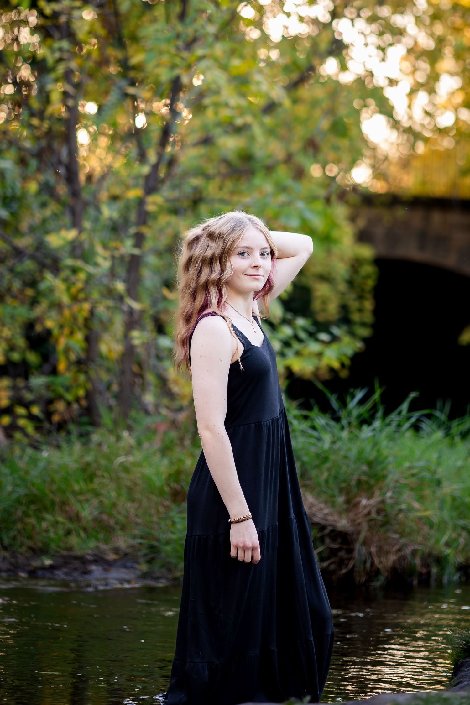 A woman in a flowing black dress poses gracefully by a pond in a natural outdoor setting during golden hour.