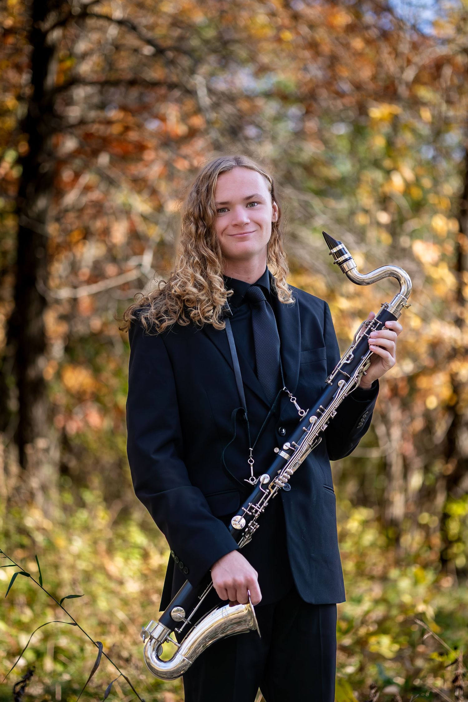A musician in a black blazer poses with a clarinet in an autumn forest setting.