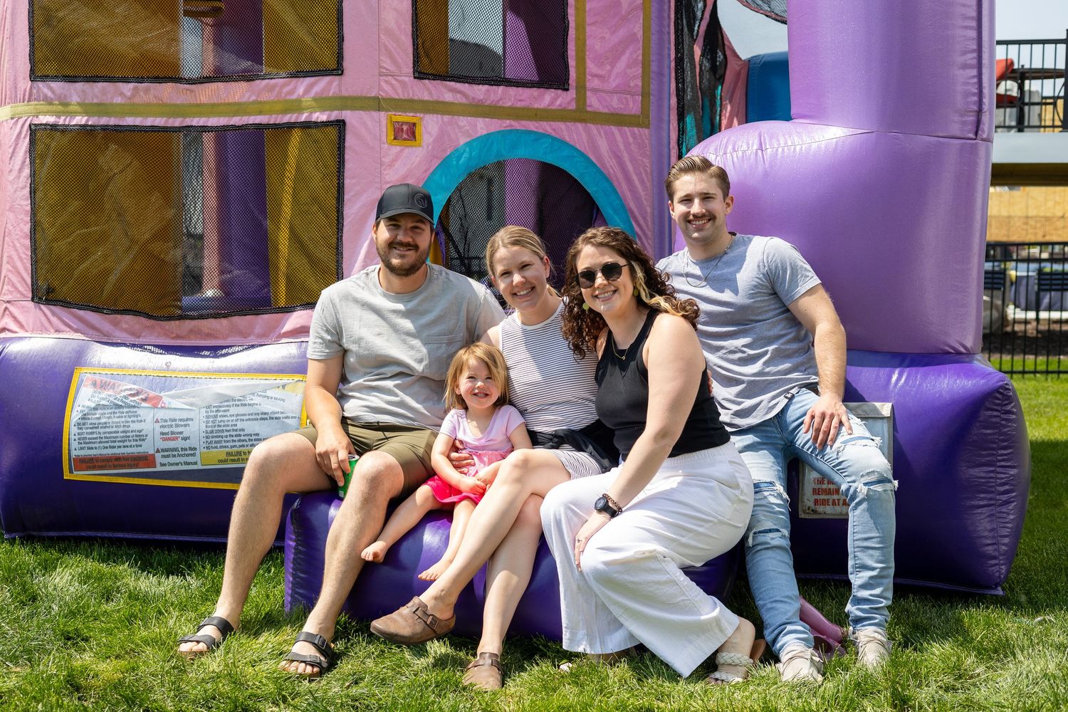 Purple bounce house castle set up on grass with people relaxing nearby.