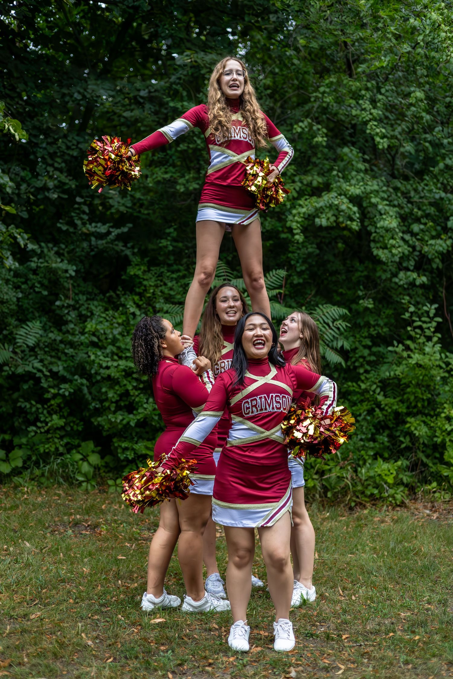 Cheerleaders perform a stunt formation outdoors while holding red and gold pom poms.