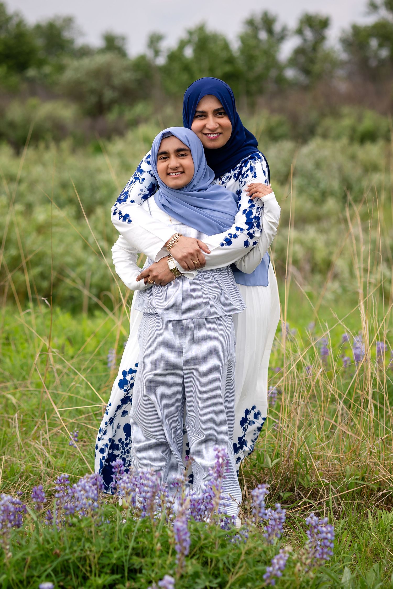 Muslim mother and daughter hugging and smiling in a field of purple wildflowers wearing hijabs.