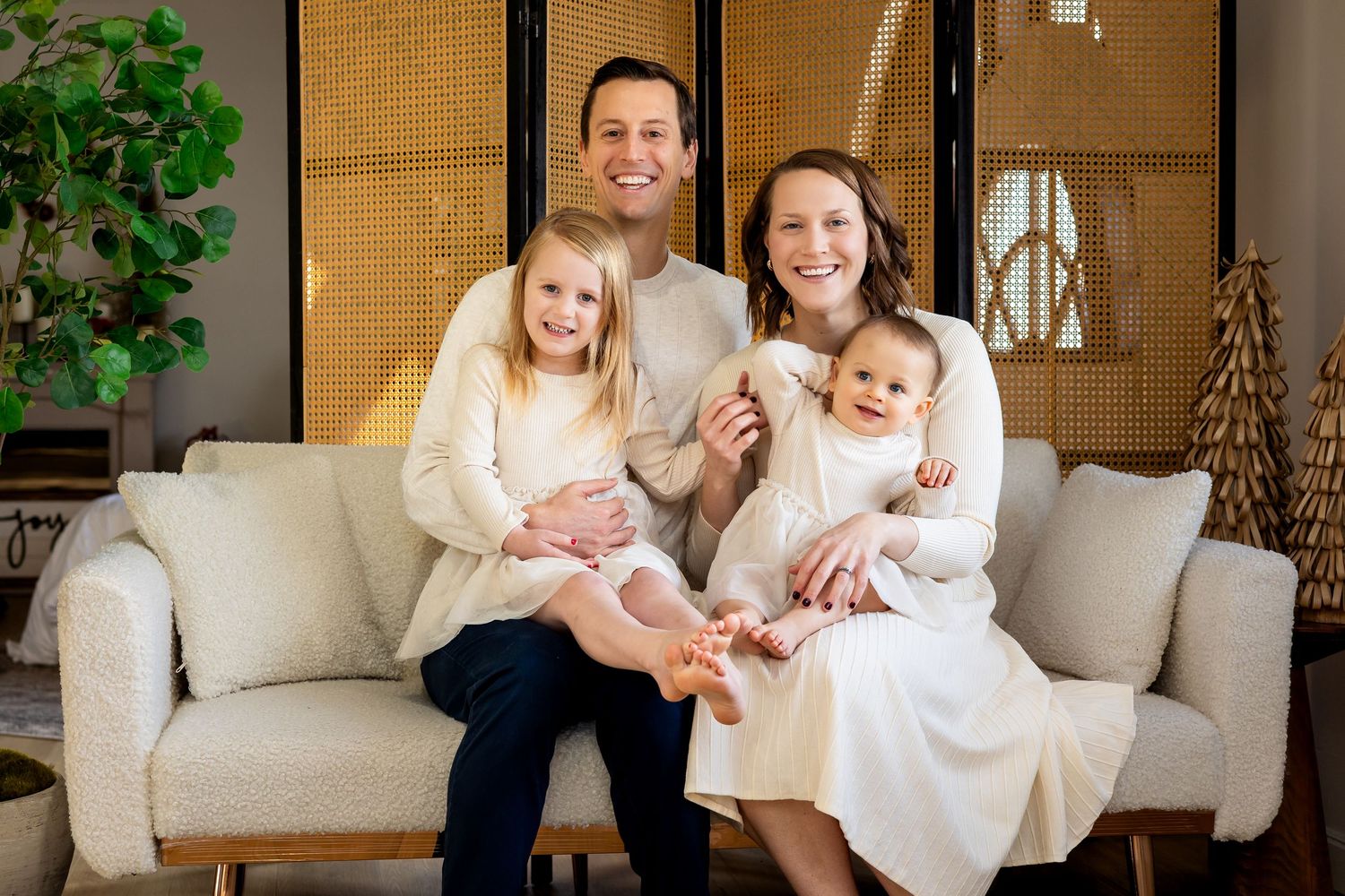 Happy family of four posing on a white sofa, parents smiling with two young children in a cozy living room.