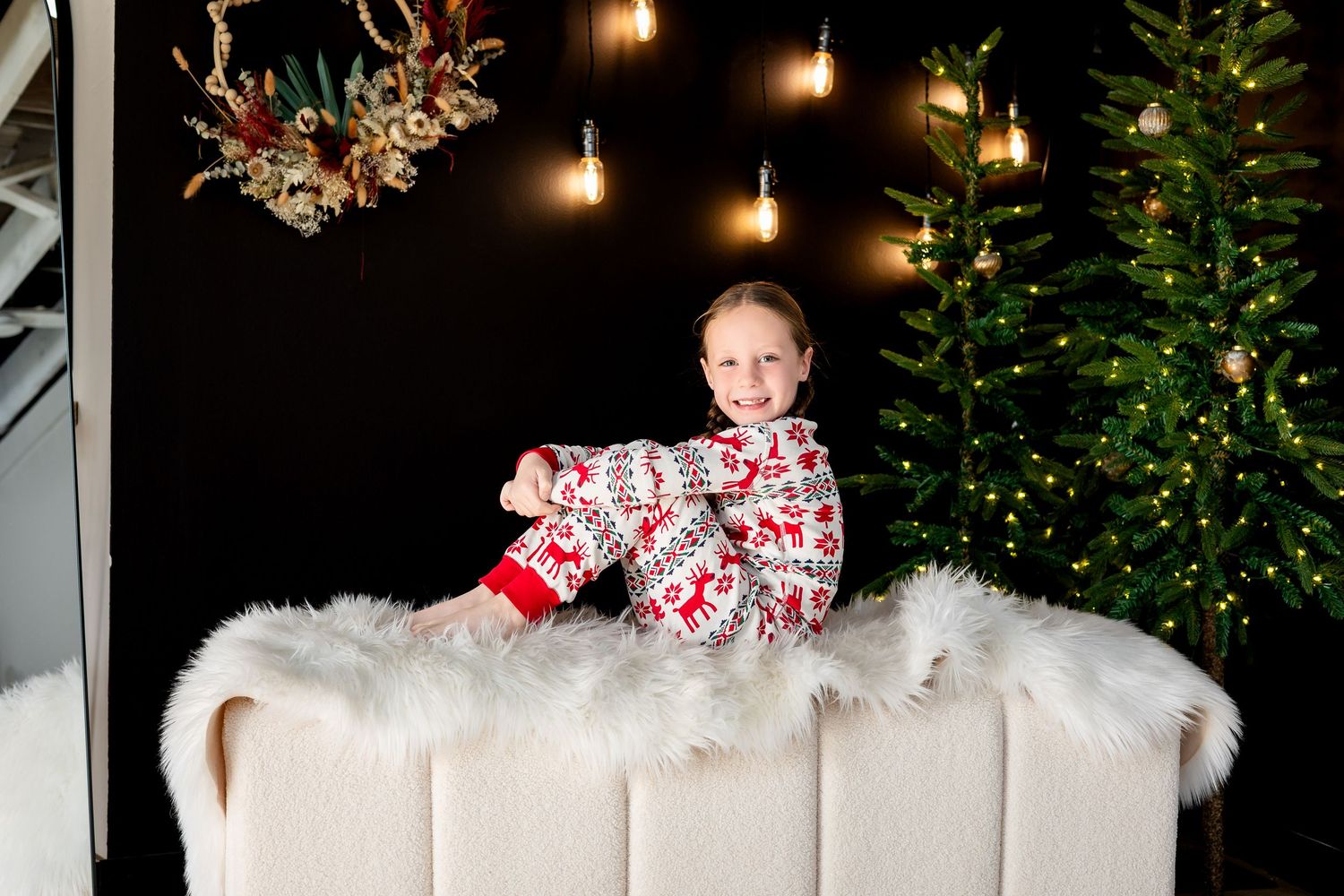 Child in Christmas pajamas sitting on a white fluffy surface near a decorated Christmas tree with festive lights.