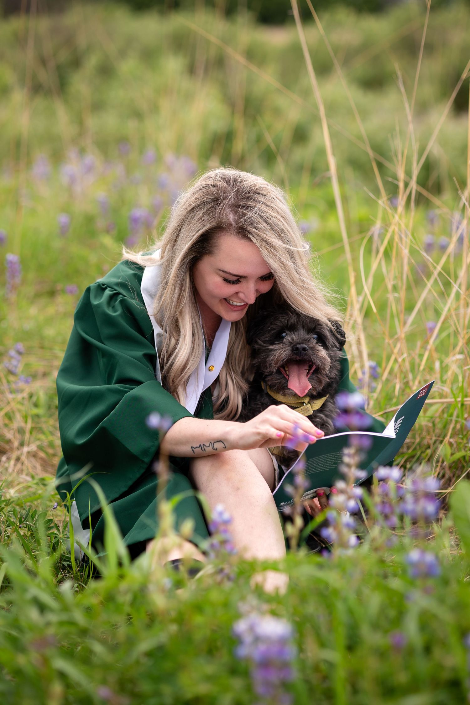 A smiling blonde woman in a green graduation gown sits in a wildflower field holding a diploma with her small black dog.