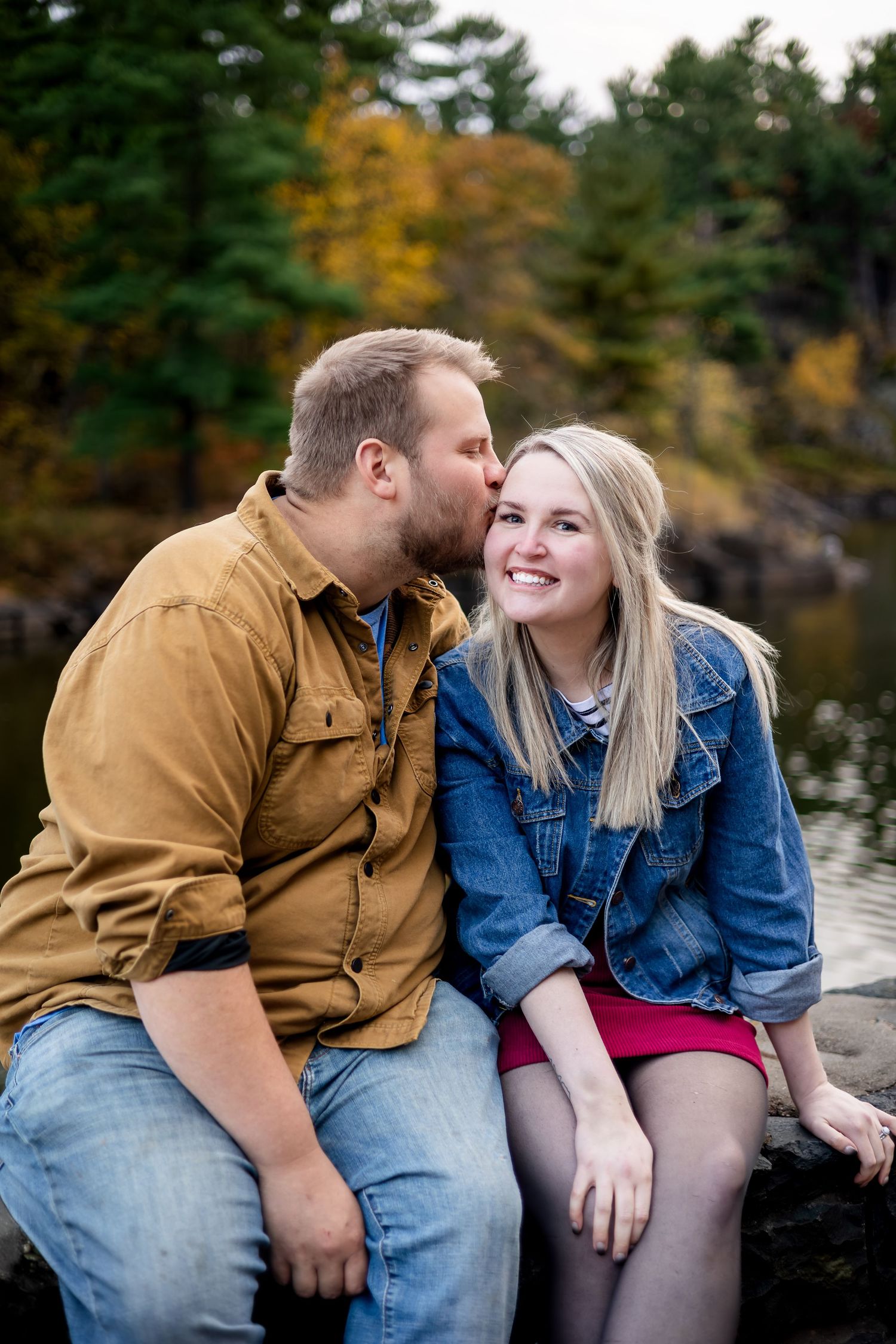 Couple sitting on rocks by autumn lake, man kissing woman on cheek as she smiles.