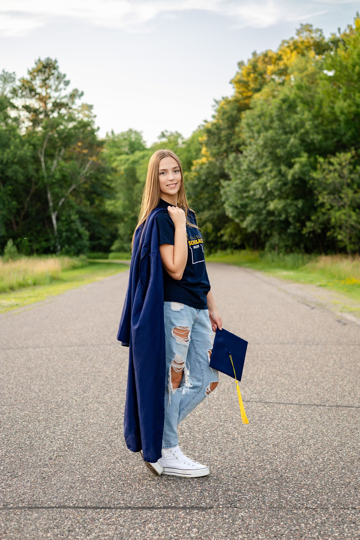 Graduate in casual attire poses with graduation cap on outdoor pathway.
