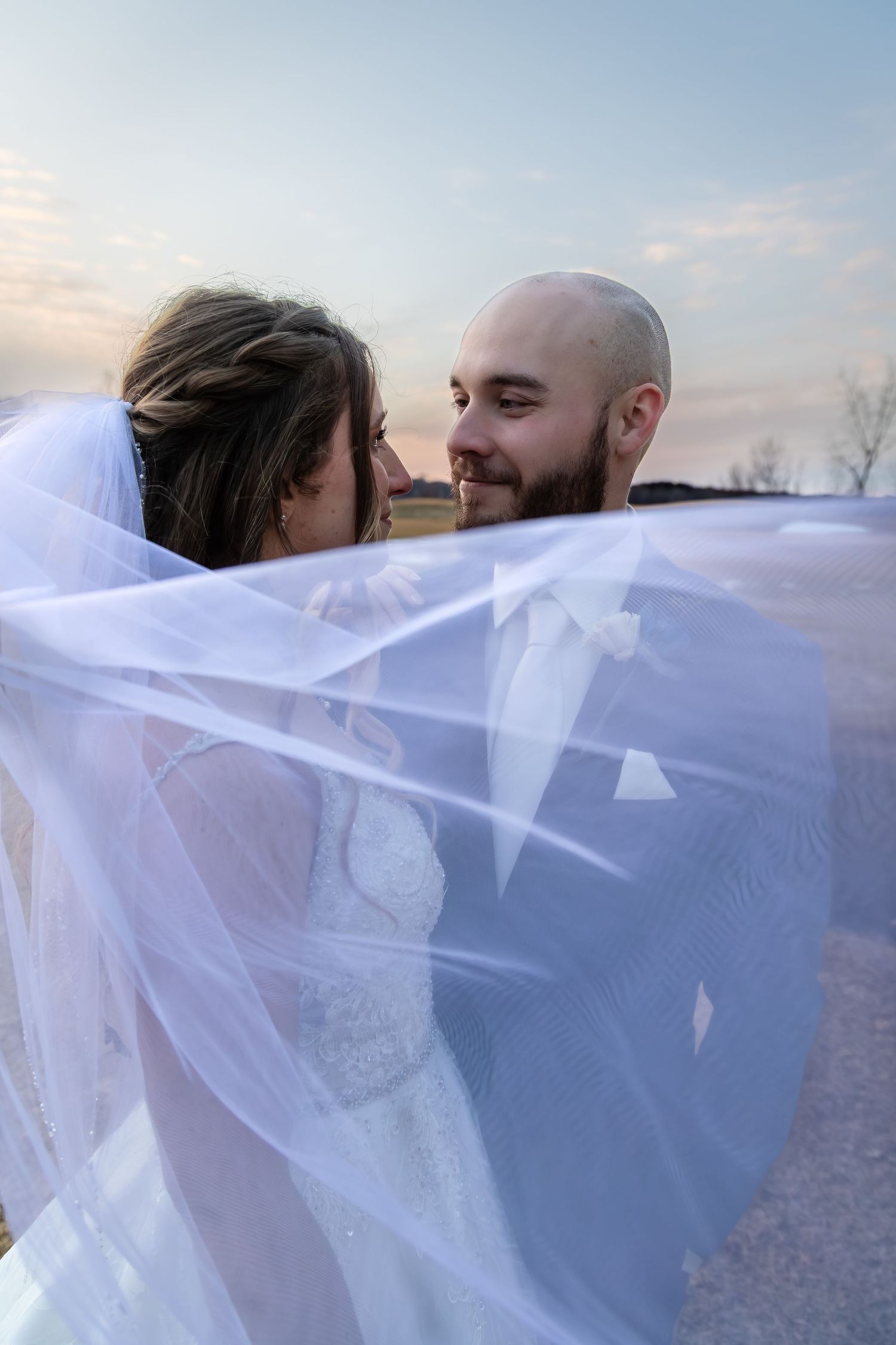 Bride and groom share a romantic moment under a flowing white veil at sunset on their wedding day.