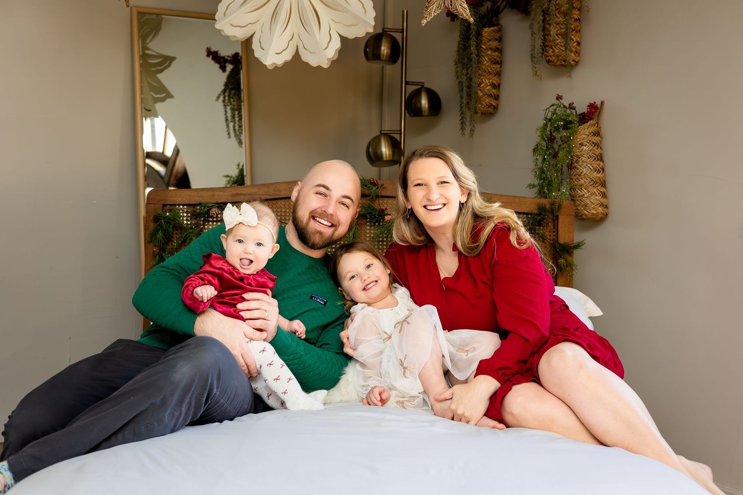 Happy family of four in holiday attire sitting on a bed, parents smiling with two young daughters in festive red and green.