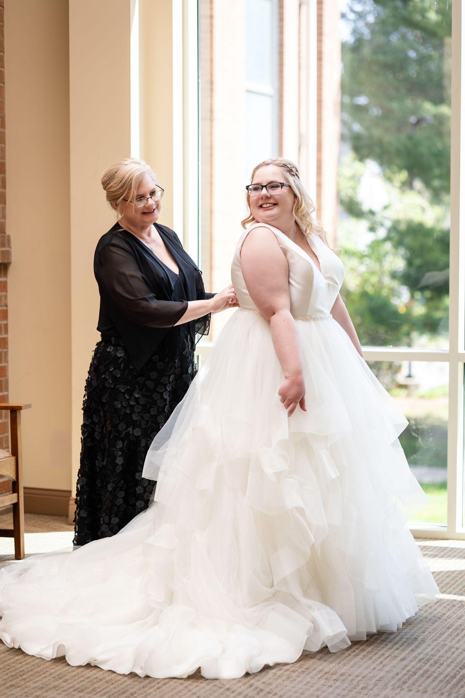 Mother helps bride in layered white tulle ball gown wedding dress near sunlit window before ceremony.