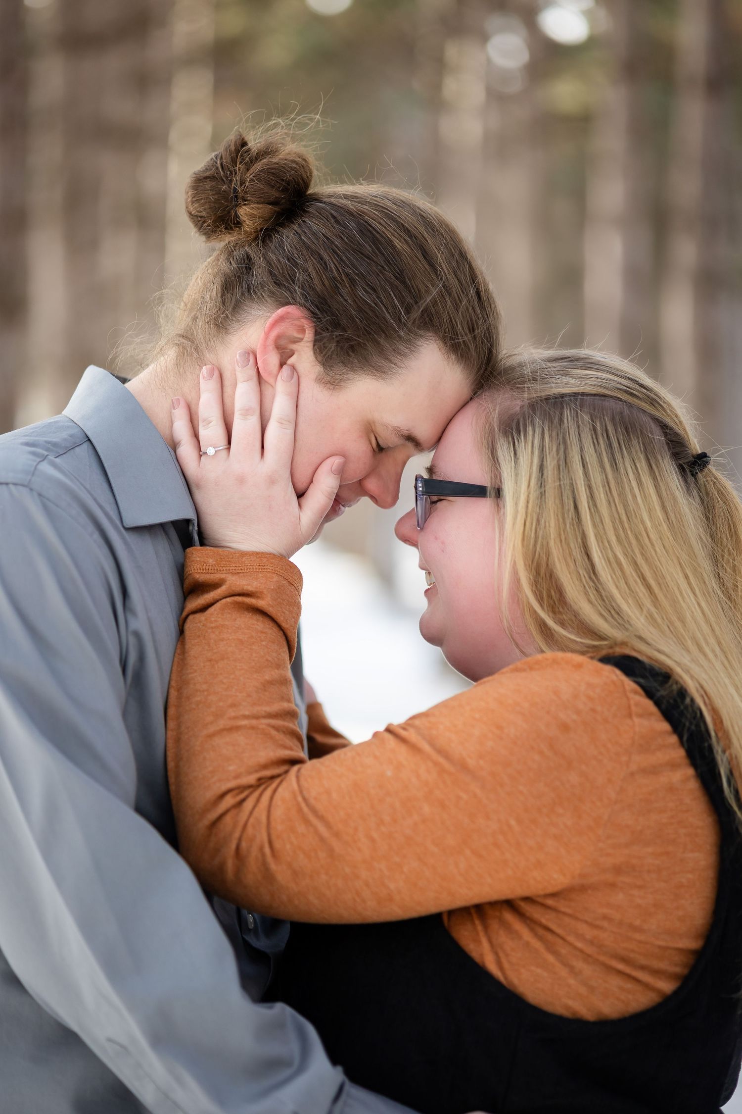 Couple sharing a tender forehead-to-forehead moment outdoors in a snowy forest, woman in glasses smiling warmly.