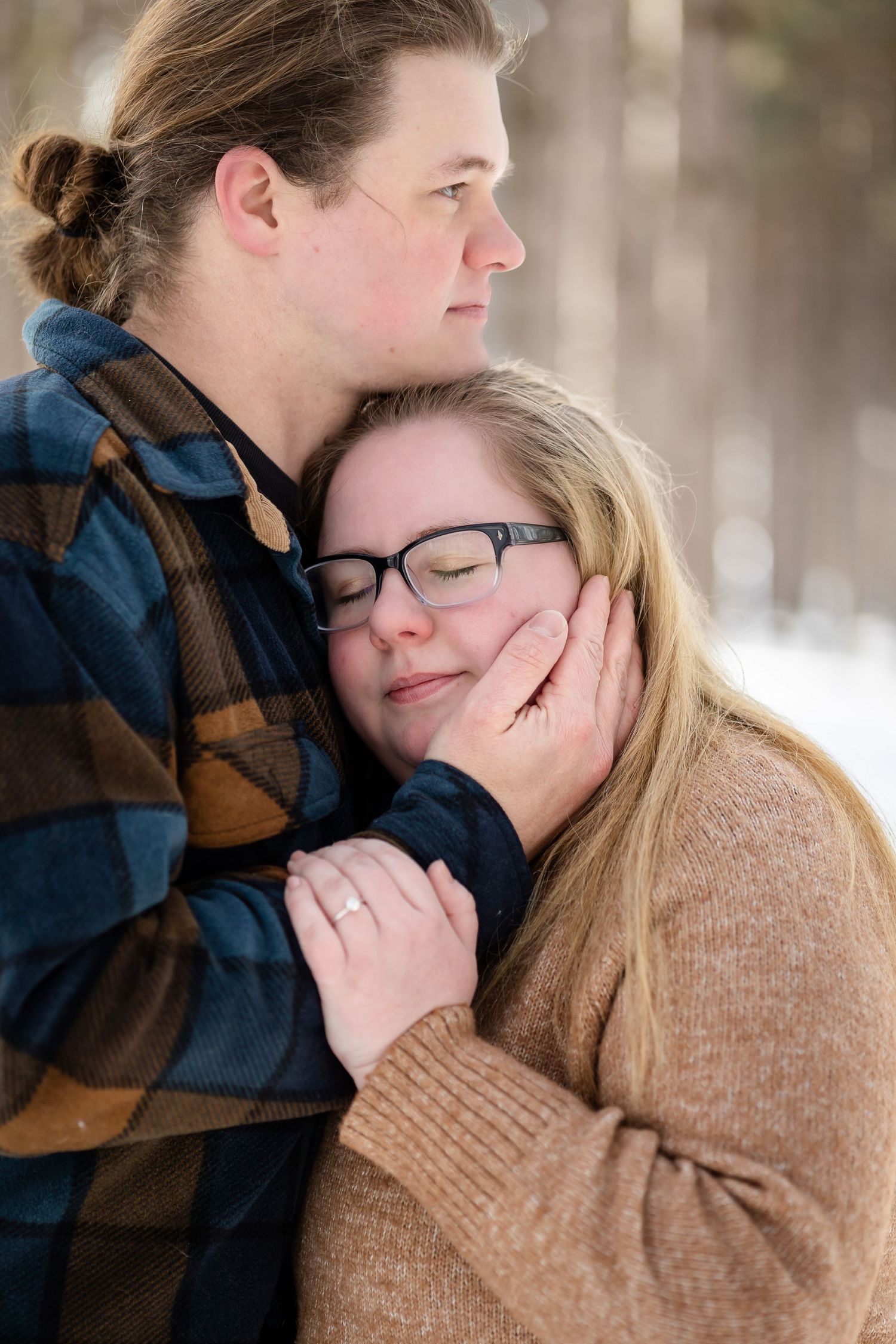 Couple embracing tenderly in snowy winter forest, woman with glasses resting head on man's chest wearing flannel shirt.