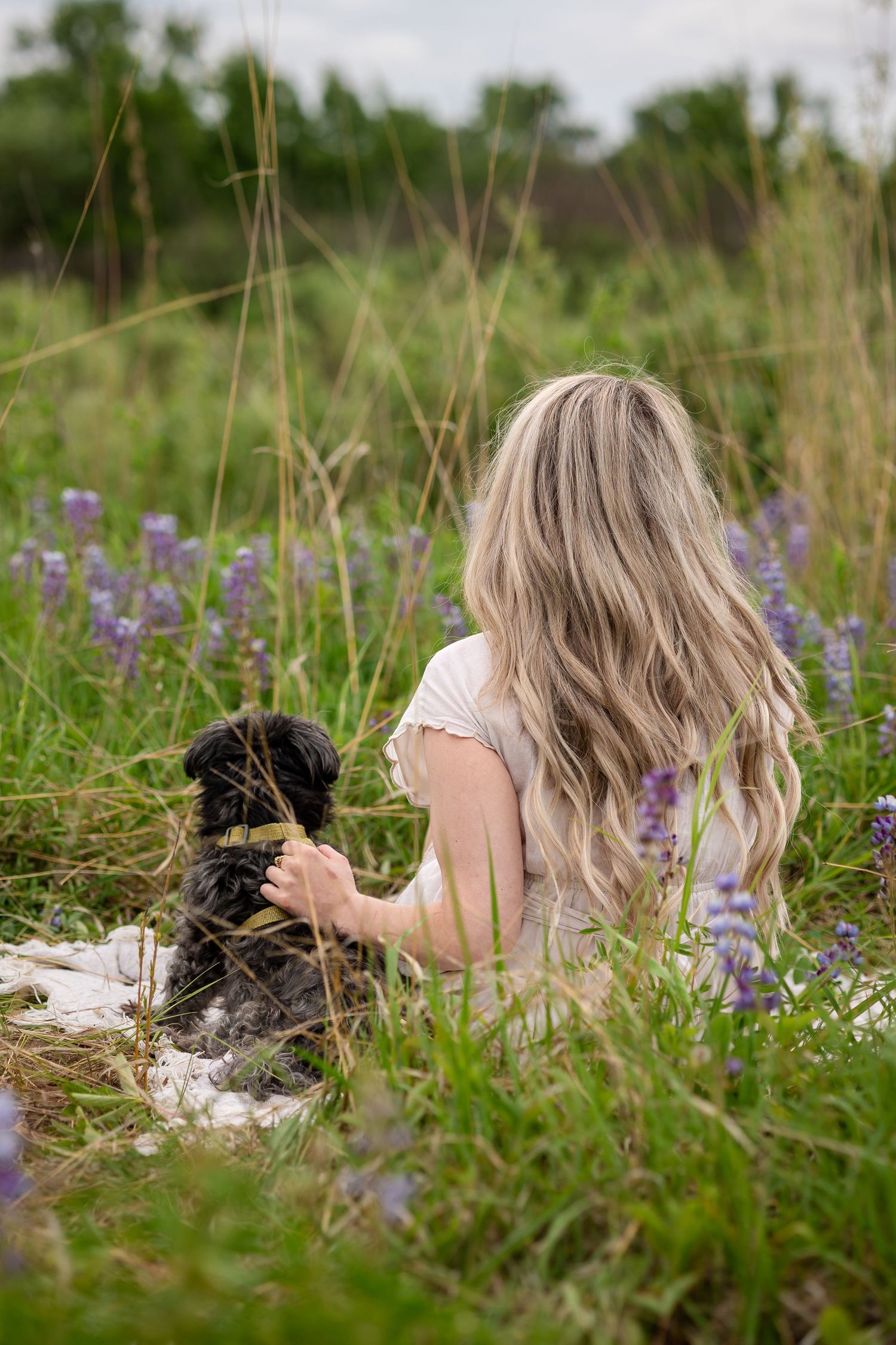 Blonde woman sitting in a field of purple wildflowers with her black dog on a cloudy day.