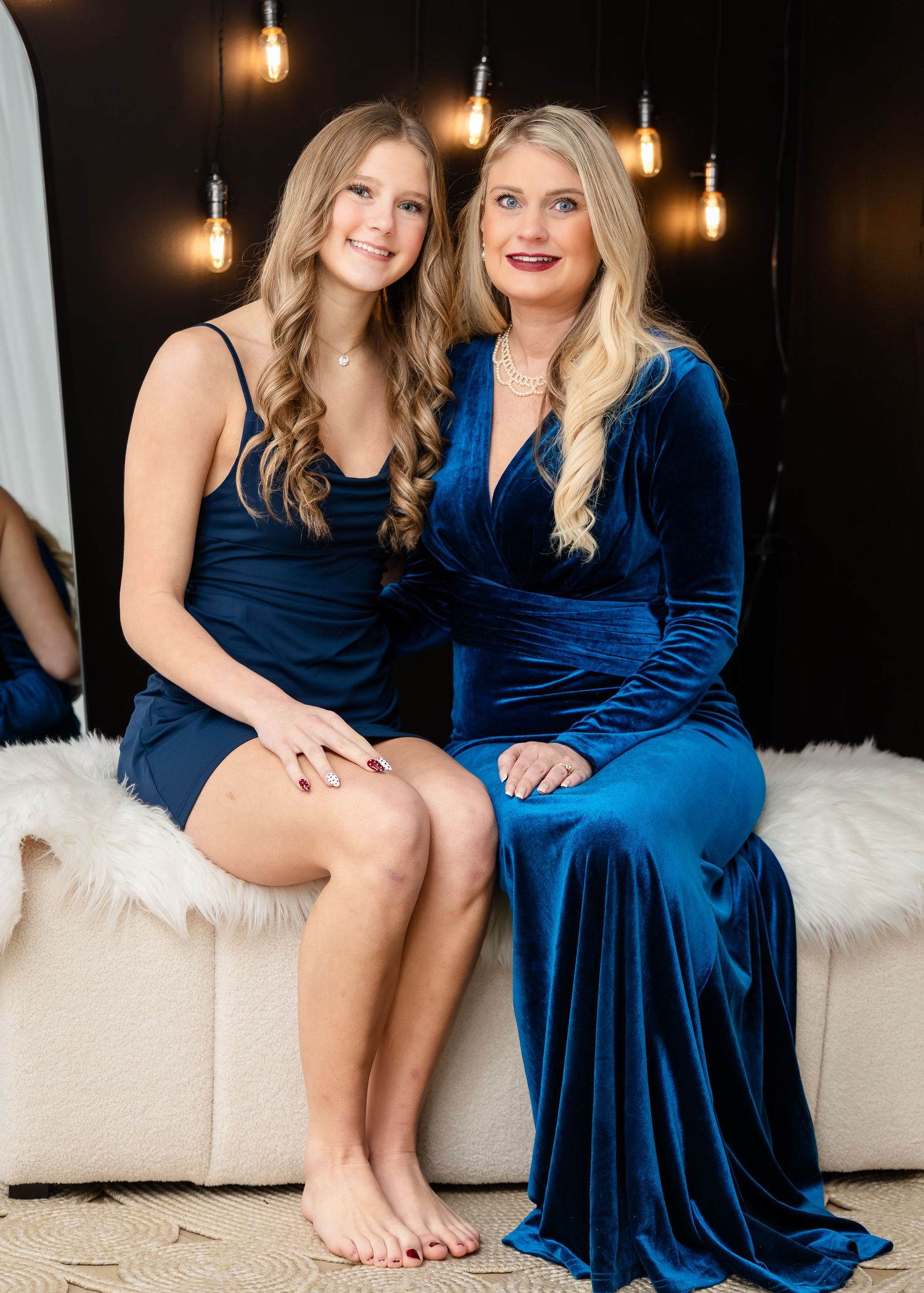 Two women in matching blue velvet dresses pose together on a white couch with warm Edison bulb lighting.