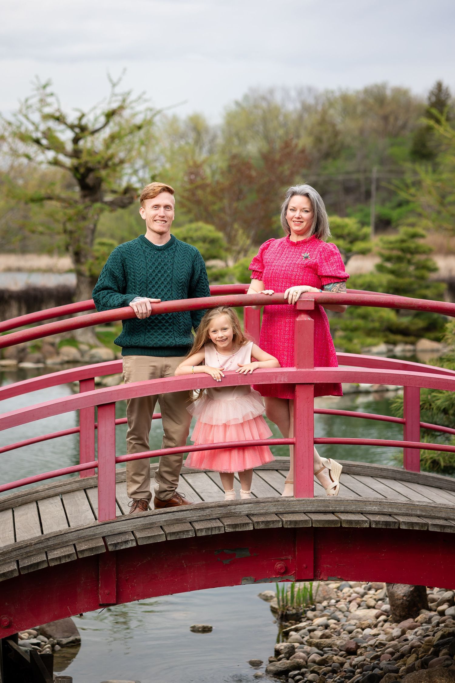 Family of three posing on a red arched bridge in a Japanese garden, girl in pink tutu dress with parents.