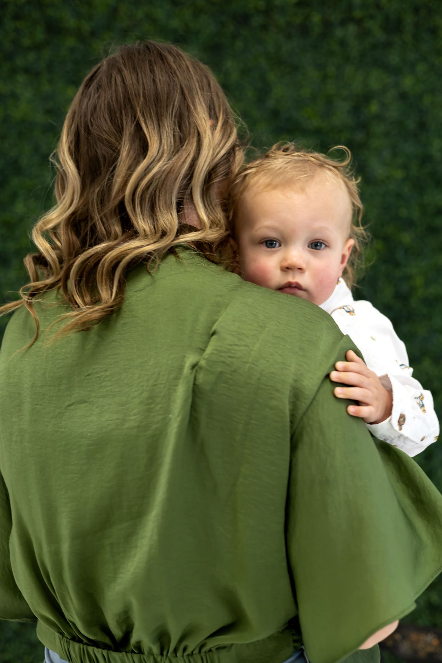 A tender moment captured as a small child peeks over a shoulder wearing a white patterned outfit.