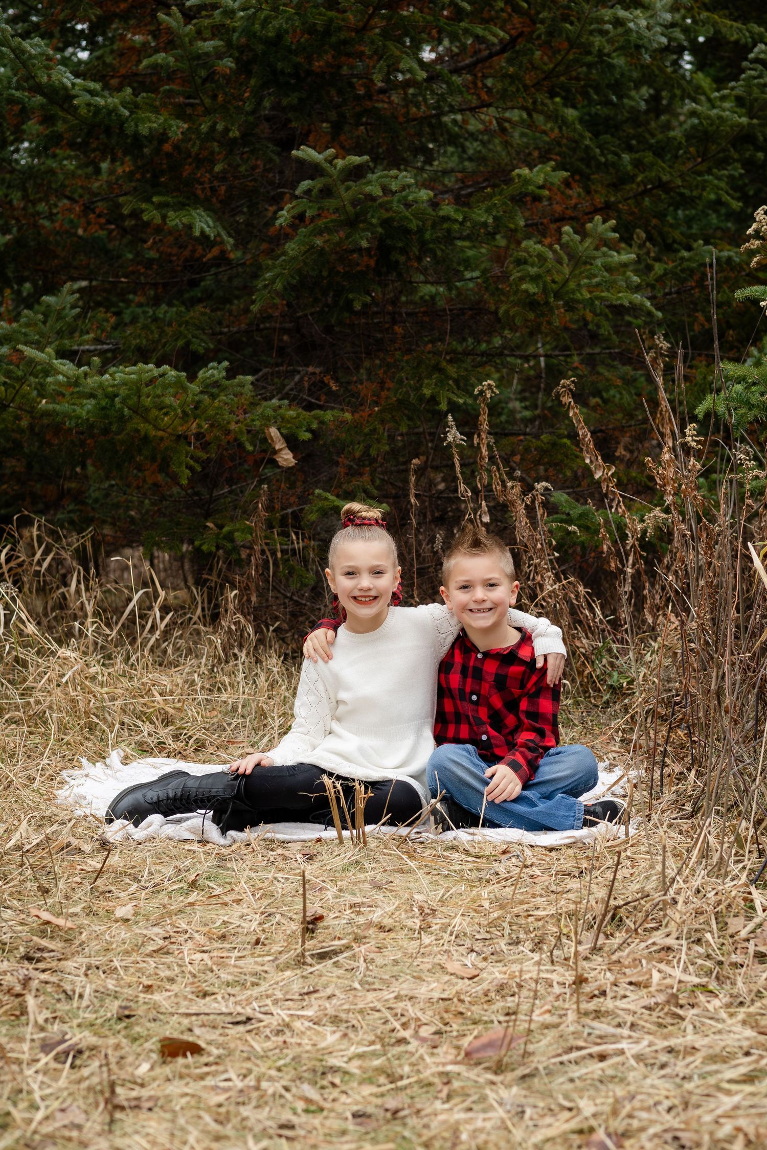 Two children sit together on a blanket in a natural outdoor setting with dried grass and trees.