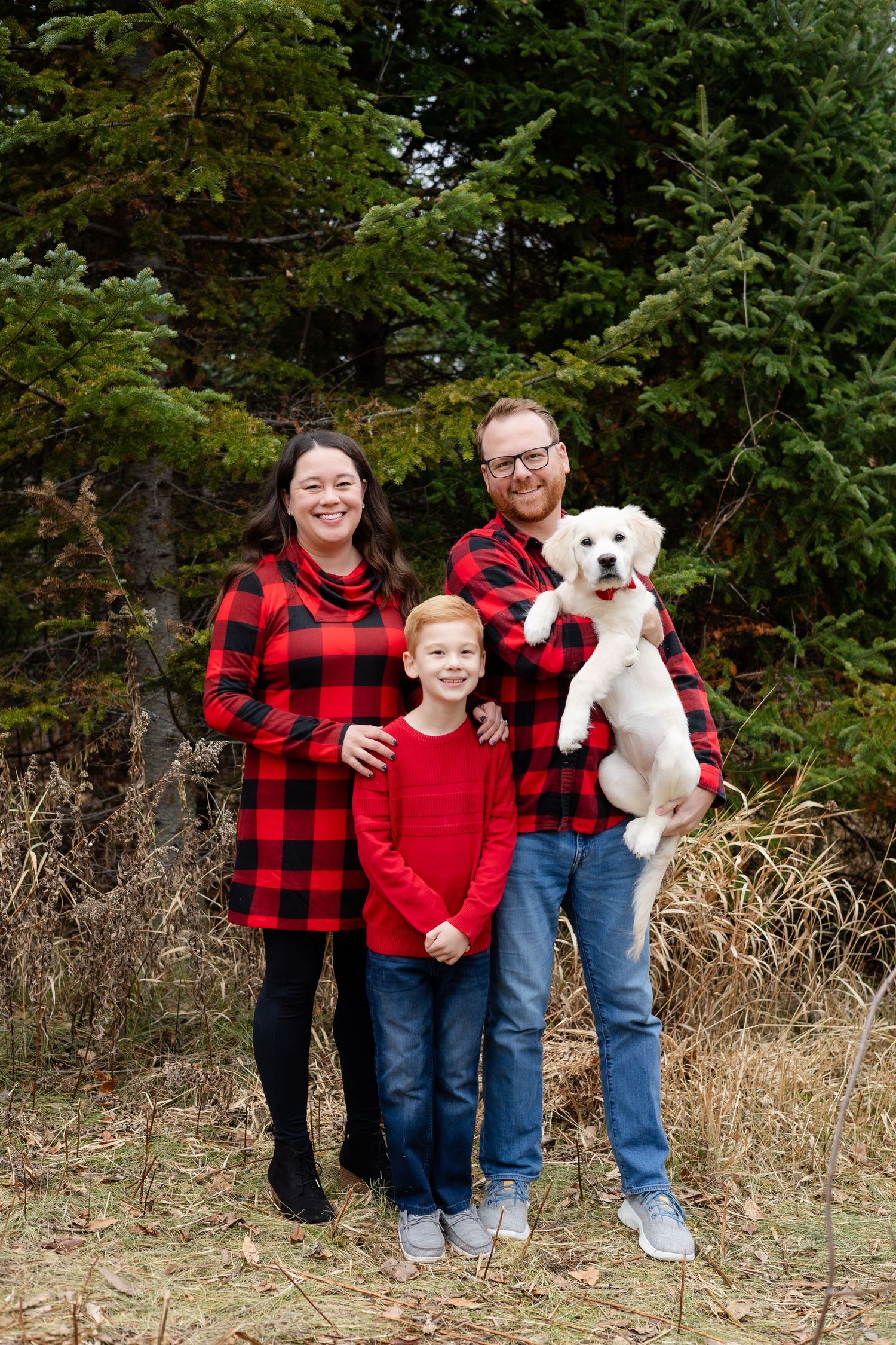 A family wearing red plaid flannel shirts poses with their white dog in a natural outdoor setting.