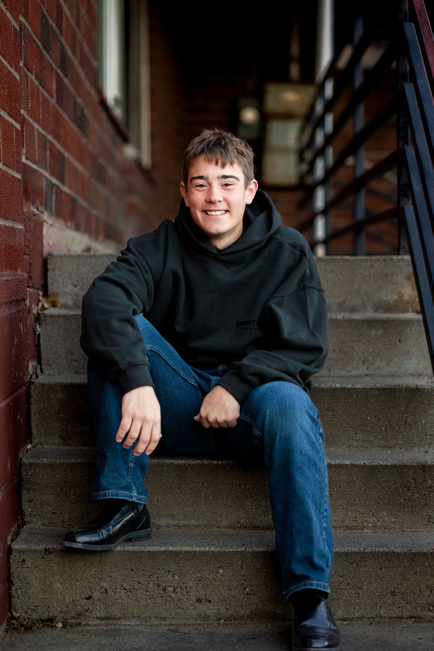 A person in a black hoodie and jeans sits casually on concrete steps against a brick wall background.