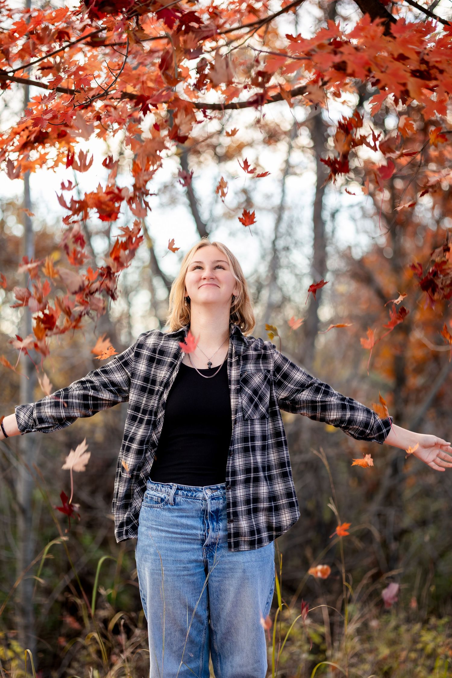 Person in plaid shirt and jeans stands with arms outstretched under vibrant red autumn maple leaves in a forest setting.