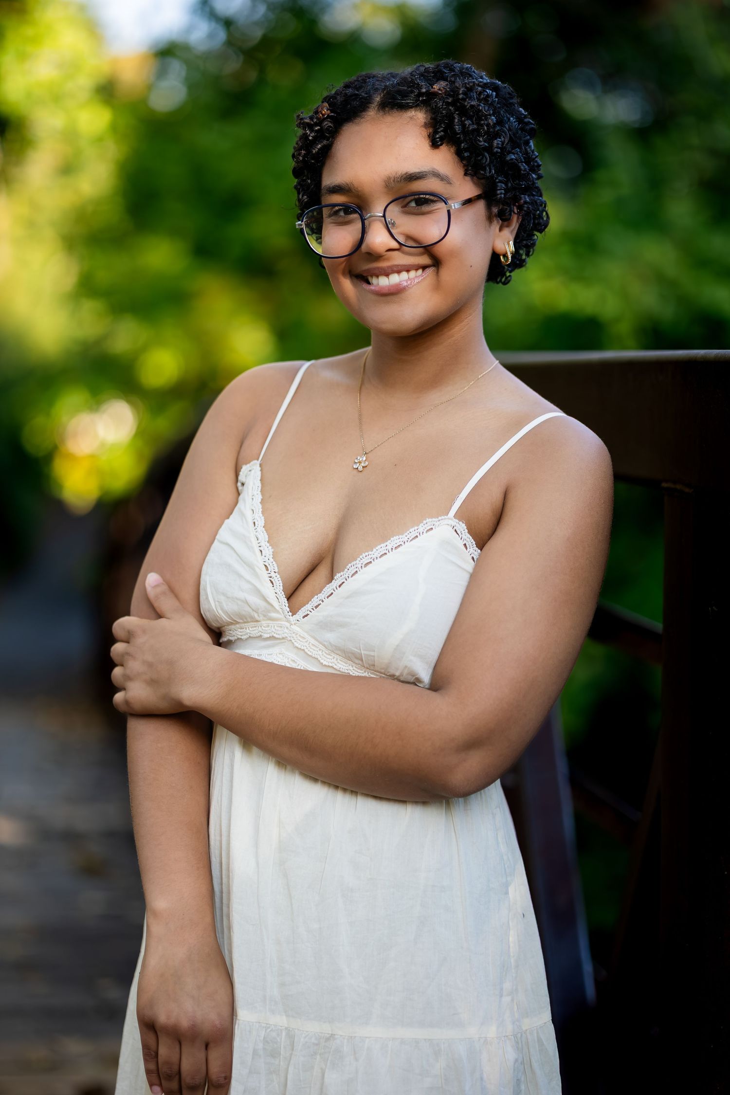 Portrait in a white lace camisole and glasses against a sunlit outdoor background with green foliage.