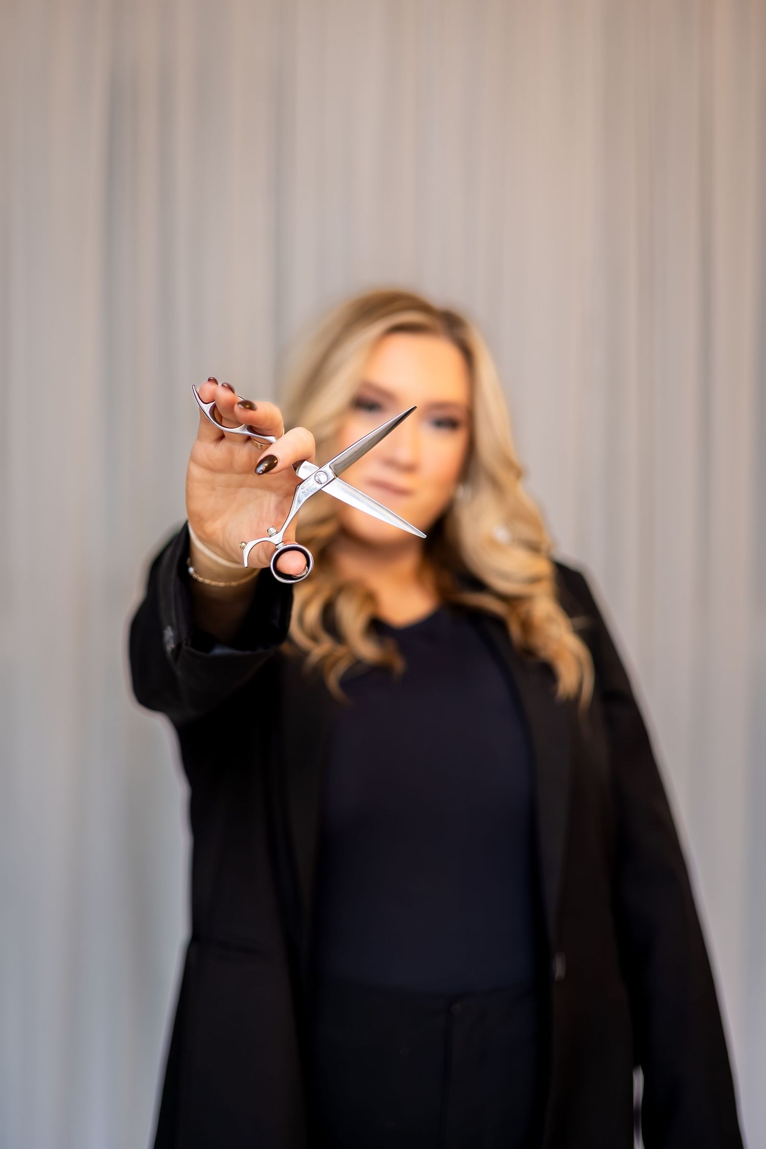 A person in a black blazer holds up scissors against a light gray curtained background.