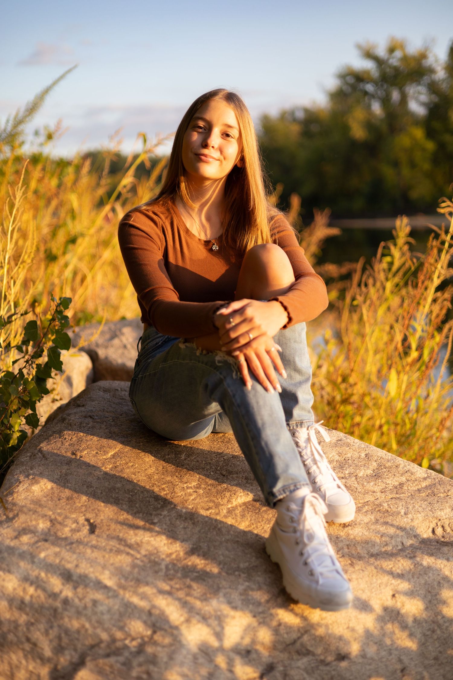 Person in brown sweater and jeans sitting on rocks during golden hour sunset.