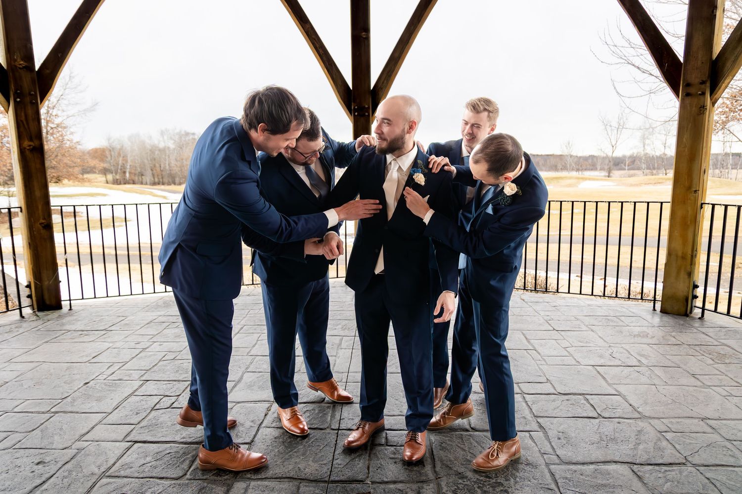 Groomsmen in navy suits adjusting the groom's boutonniere under a wooden pavilion at a wedding.