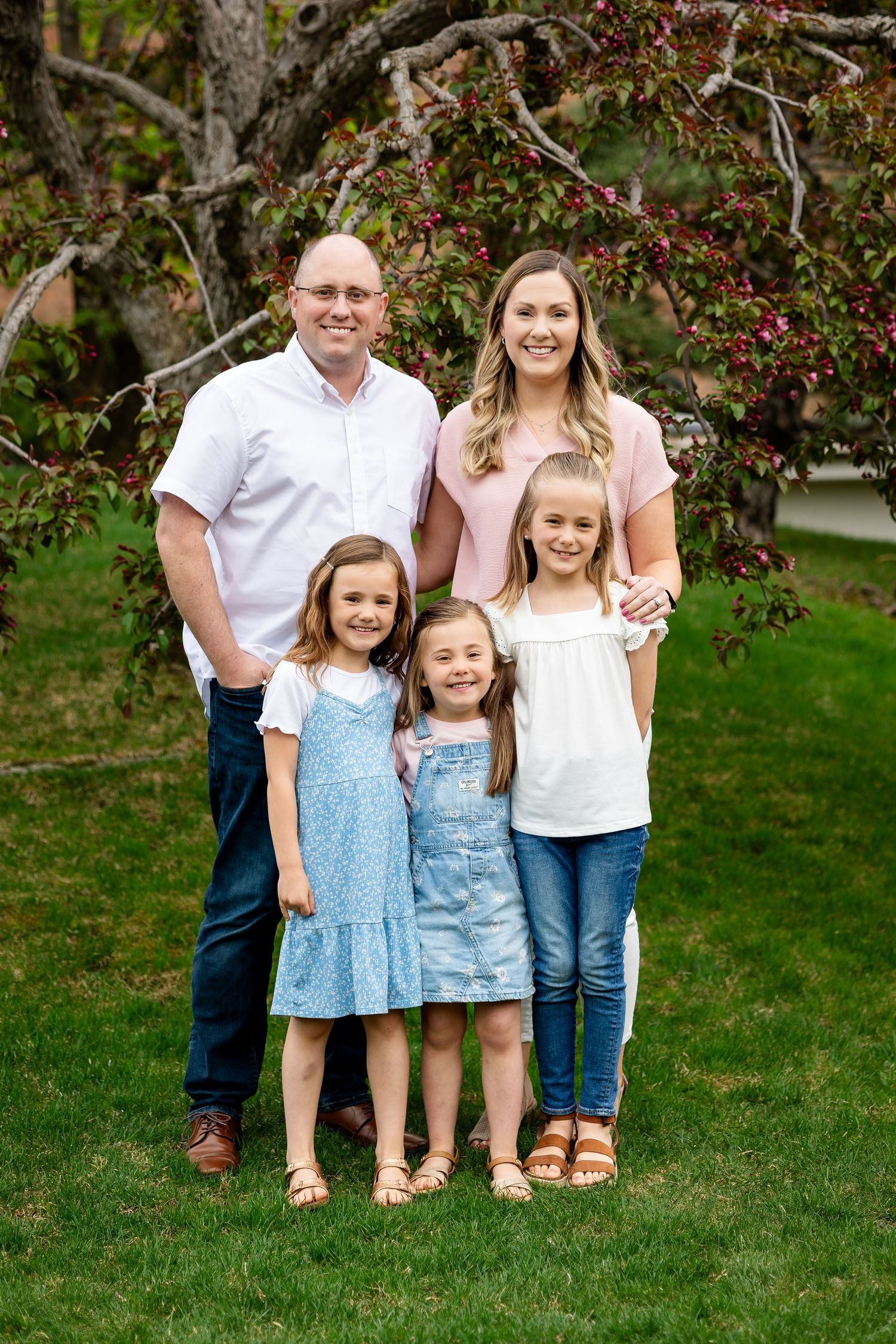 Family of five posing outdoors under a blooming tree on green grass, parents with three young daughters in coordinating outfits.