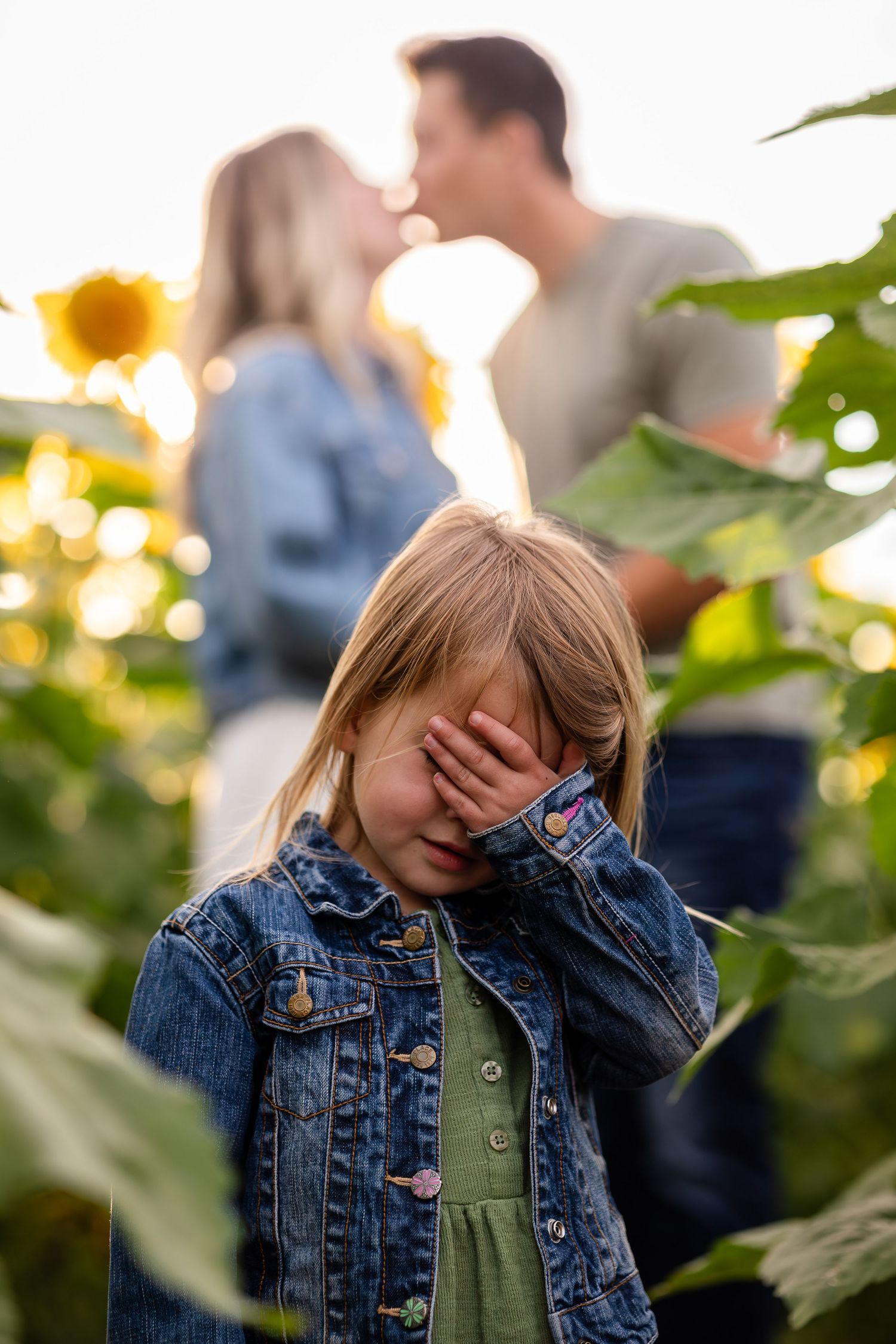 Child in denim jacket playing peek-a-boo in a sunflower field.