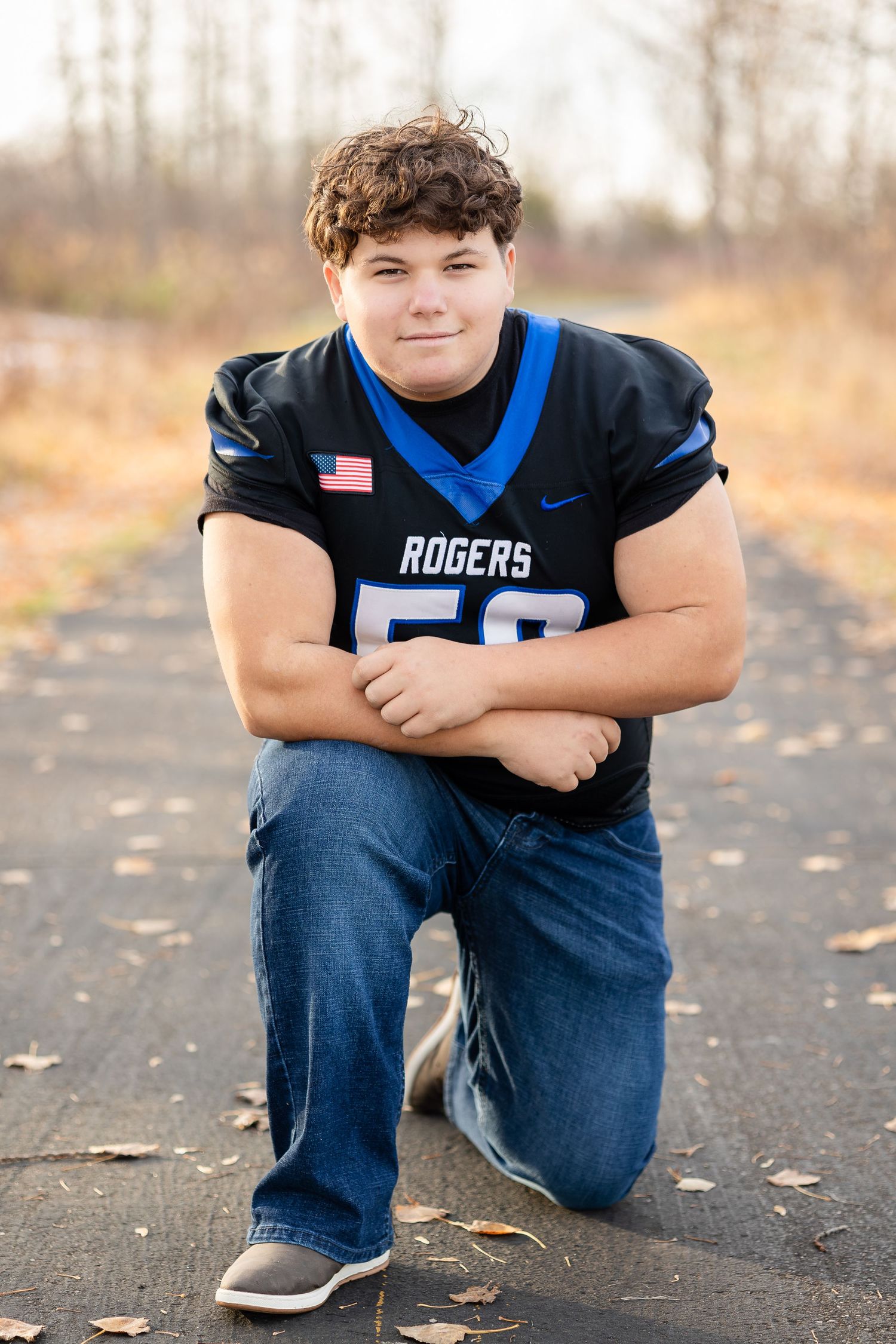 Person in Rogers football uniform number 50 kneeling on dark pavement with autumn leaves.