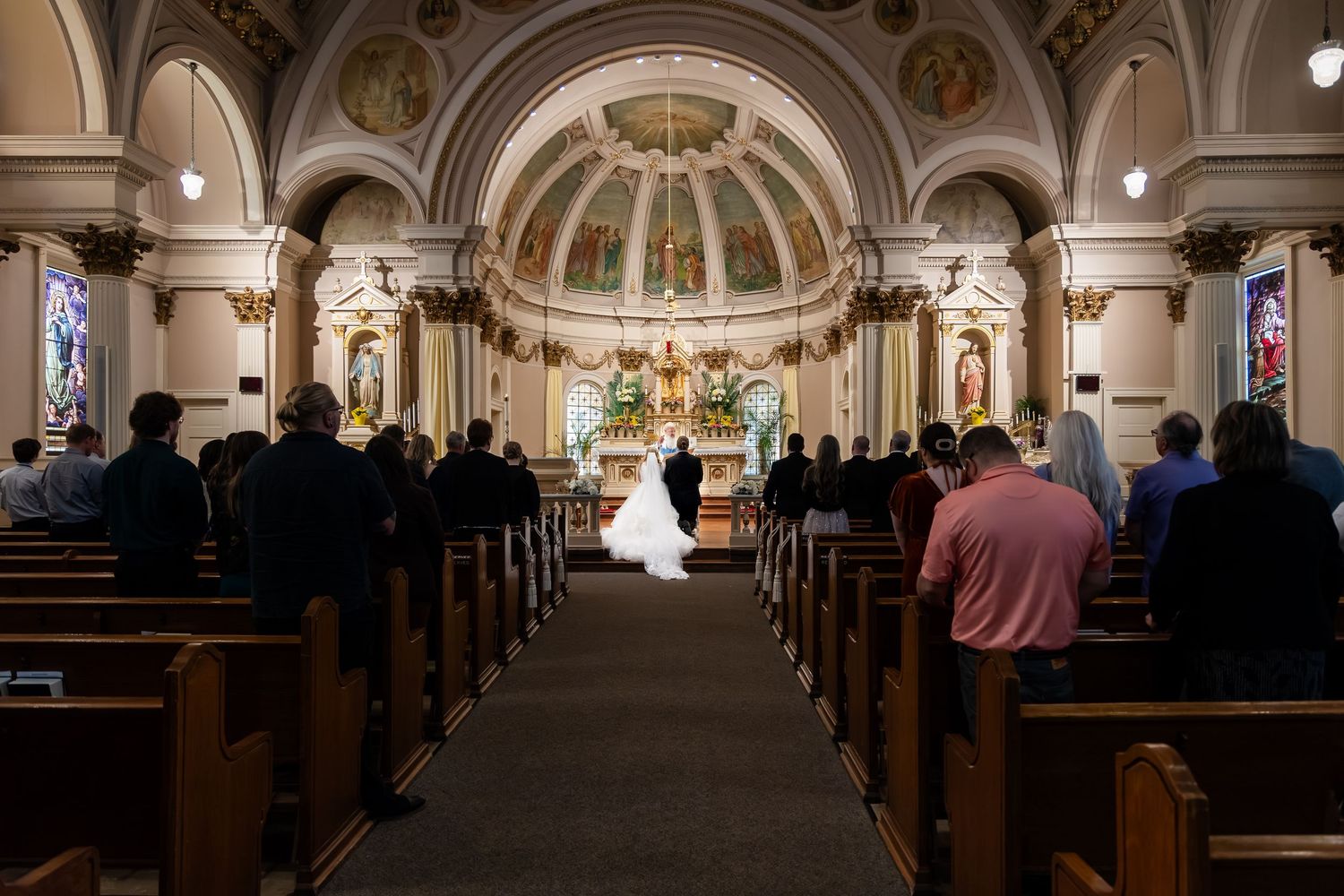 Guests stand in wooden pews as bride and groom exchange vows at ornate church altar.