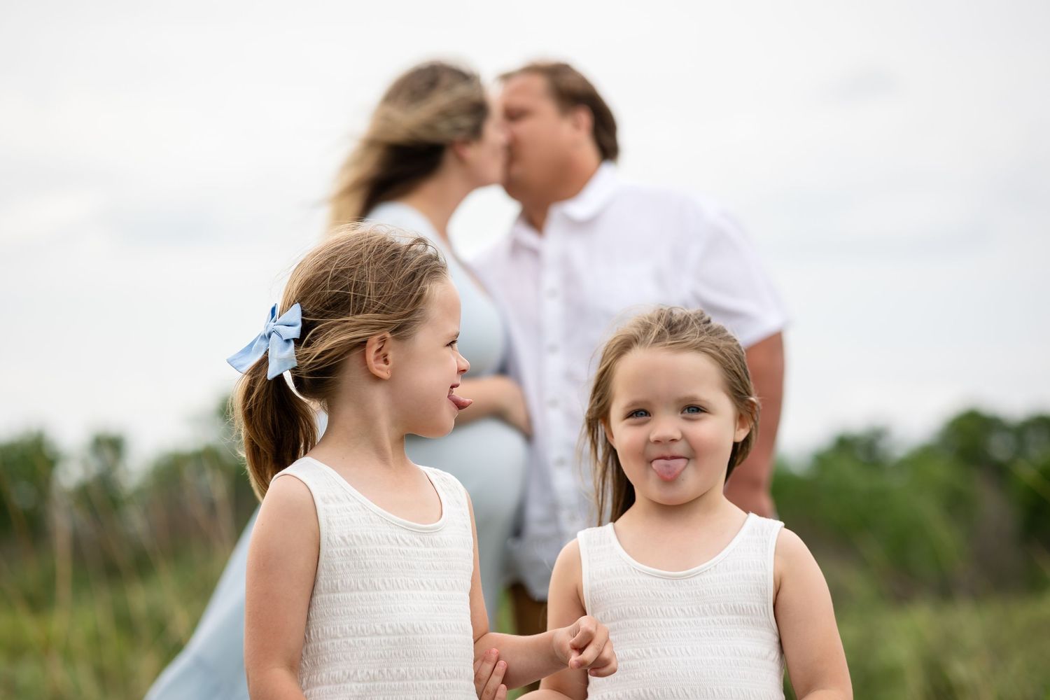 Two young girls in white tank tops smile outdoors while their parents share a kiss in the background.