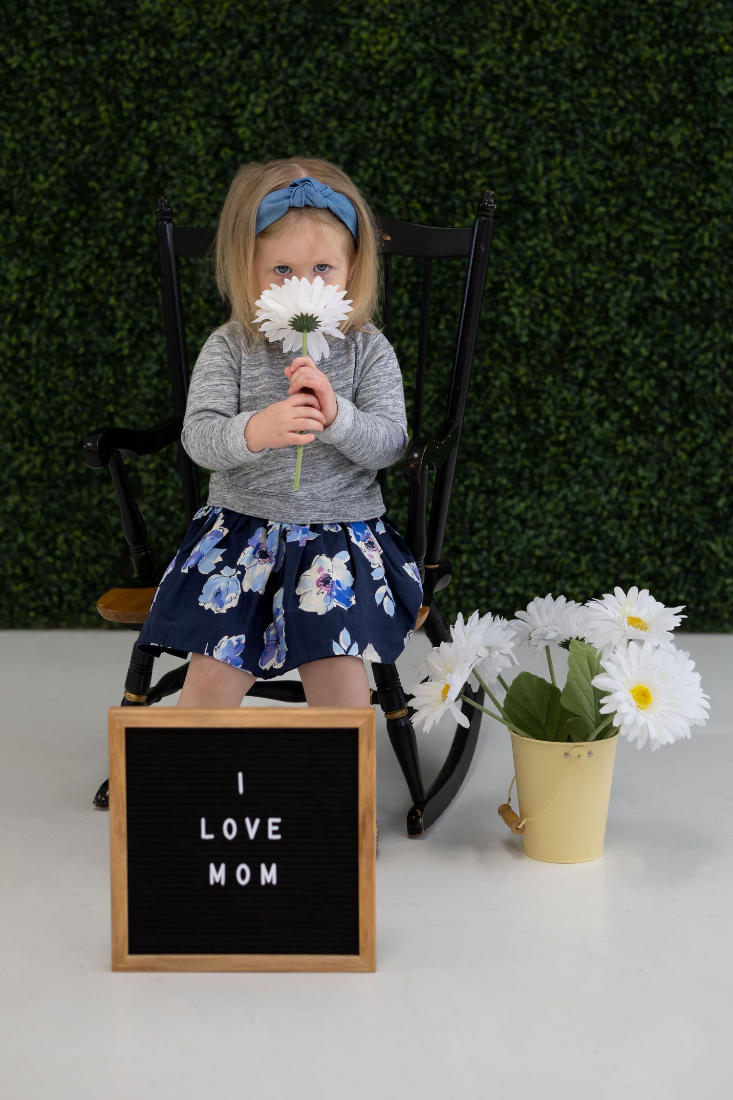A child in a floral dress holds white daisies while sitting in a rocking chair with an 'I Love Mom' sign.